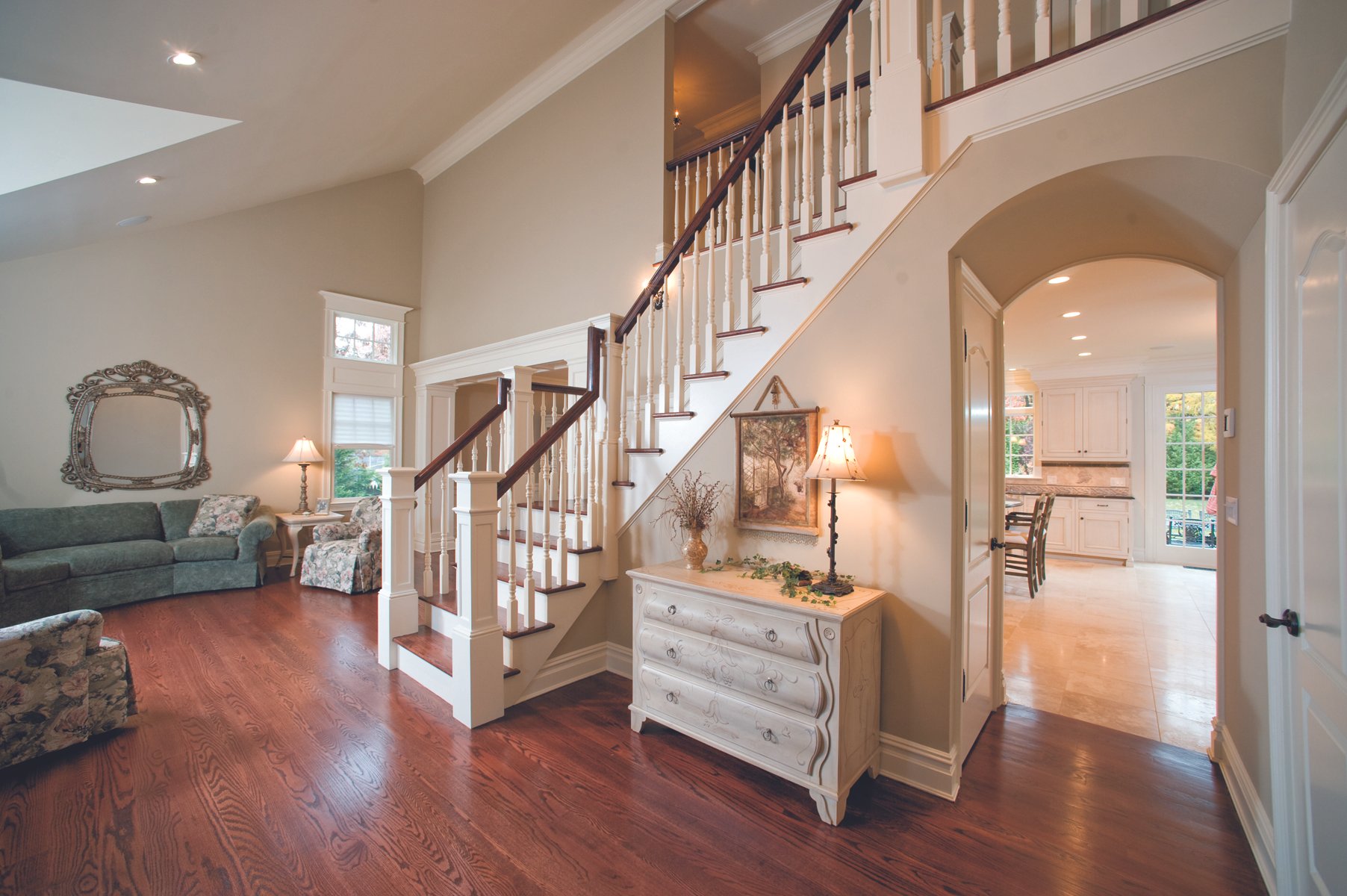 Interior of a spacious house featuring a staircase with white spindles and dark wooden handrails, a white dresser with a table lamp, a floral armchair, a sofa, and an open kitchen with white cabinetry.