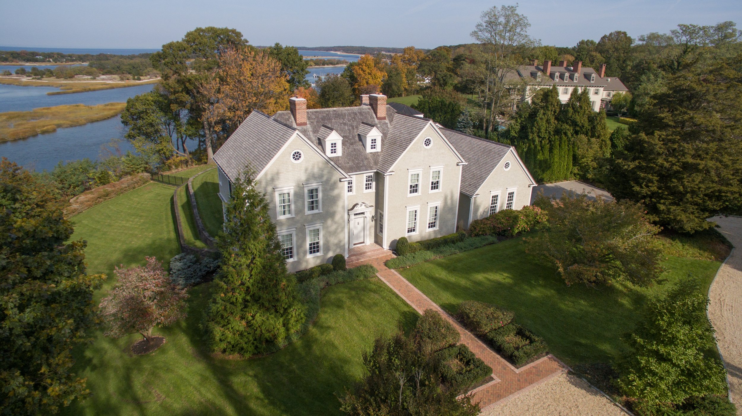 A large, elegant white house with multiple chimneys and a brick pathway leading to the front door, surrounded by manicured lawns, trees, and overlooking a river or lake in the background.