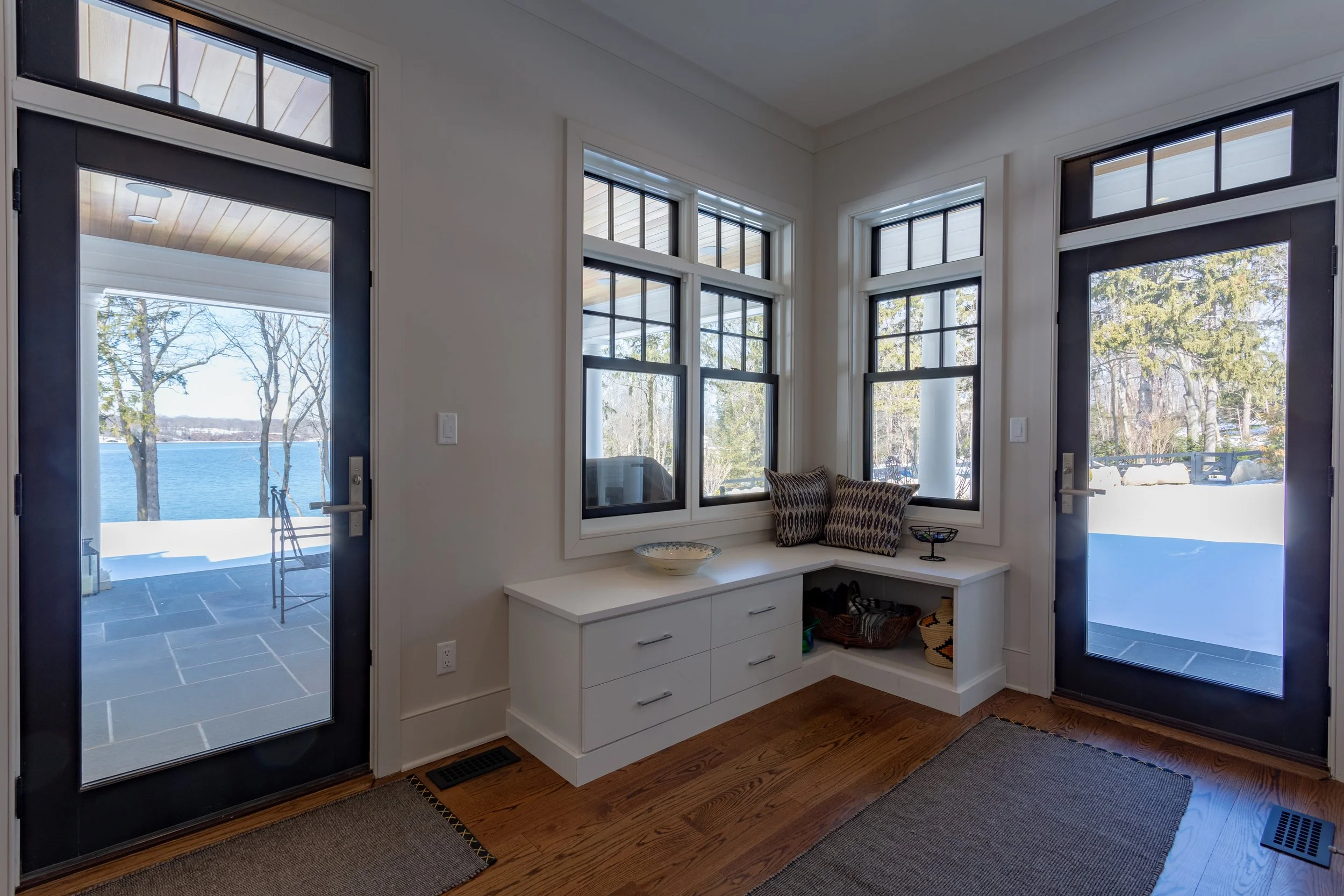 Interior view of a cozy porch with white walls, hardwood flooring, a built-in white bench with drawers, and large windows with black frames, overlooking a snowy landscape and a lake.