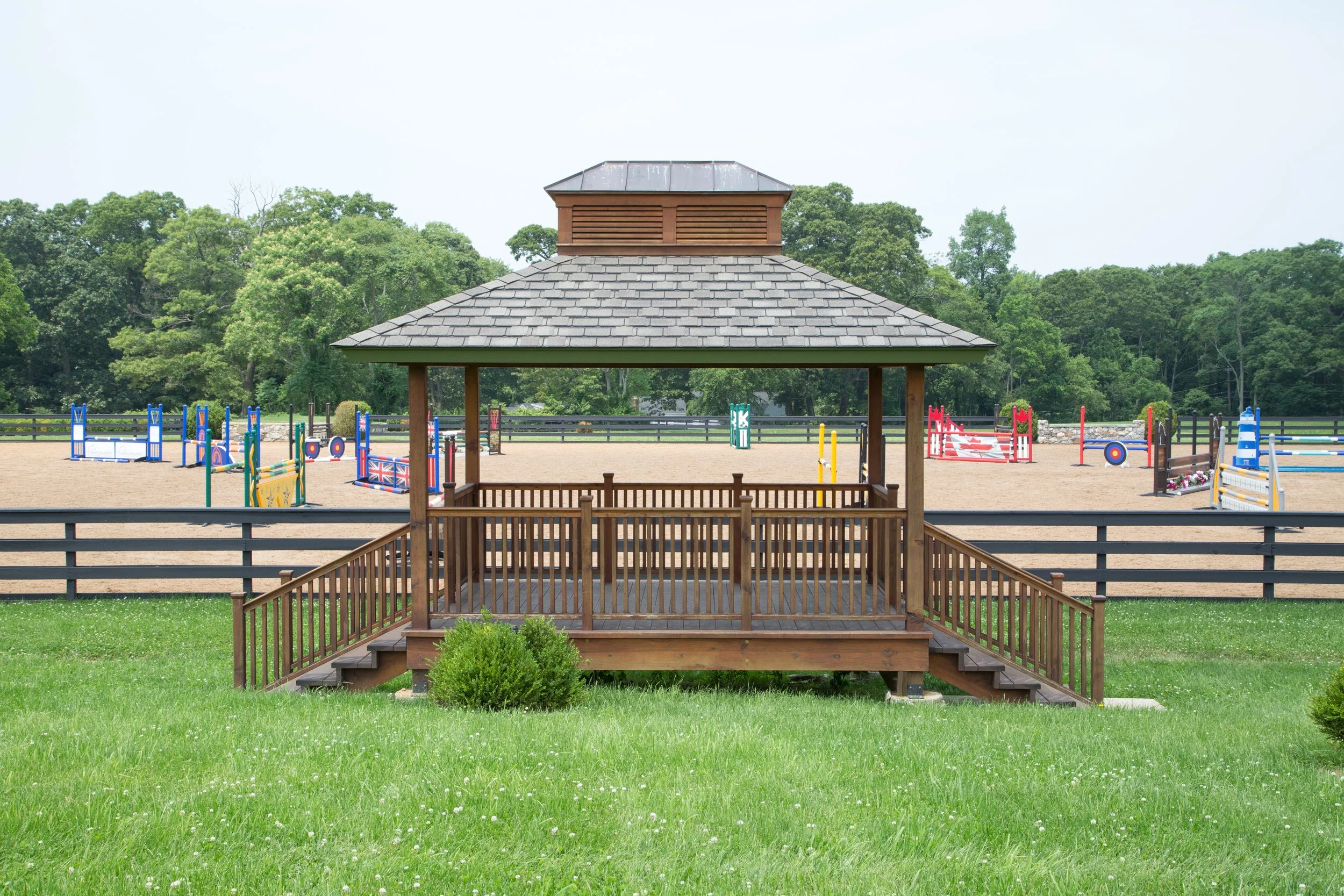 Wooden gazebo overlooking an outdoor equestrian jumping arena with colorful jumps, surrounded by green trees.