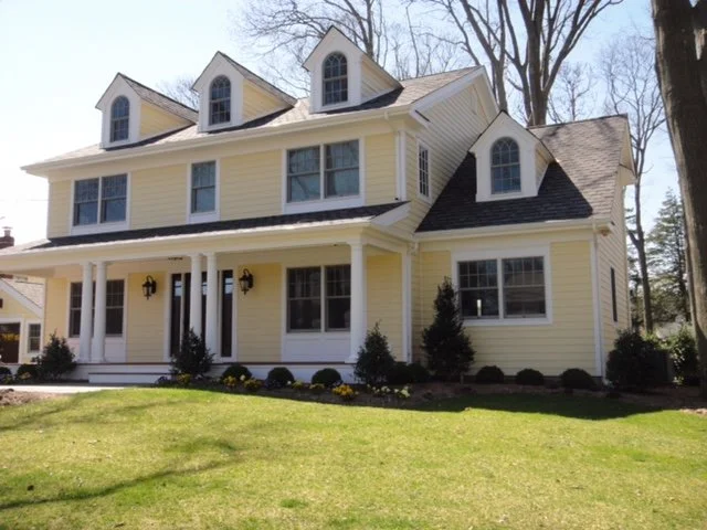 A yellow two-story house with white trim and multiple gabled dormer windows, front porch with white pillars, surrounded by a green lawn and trees in the background.