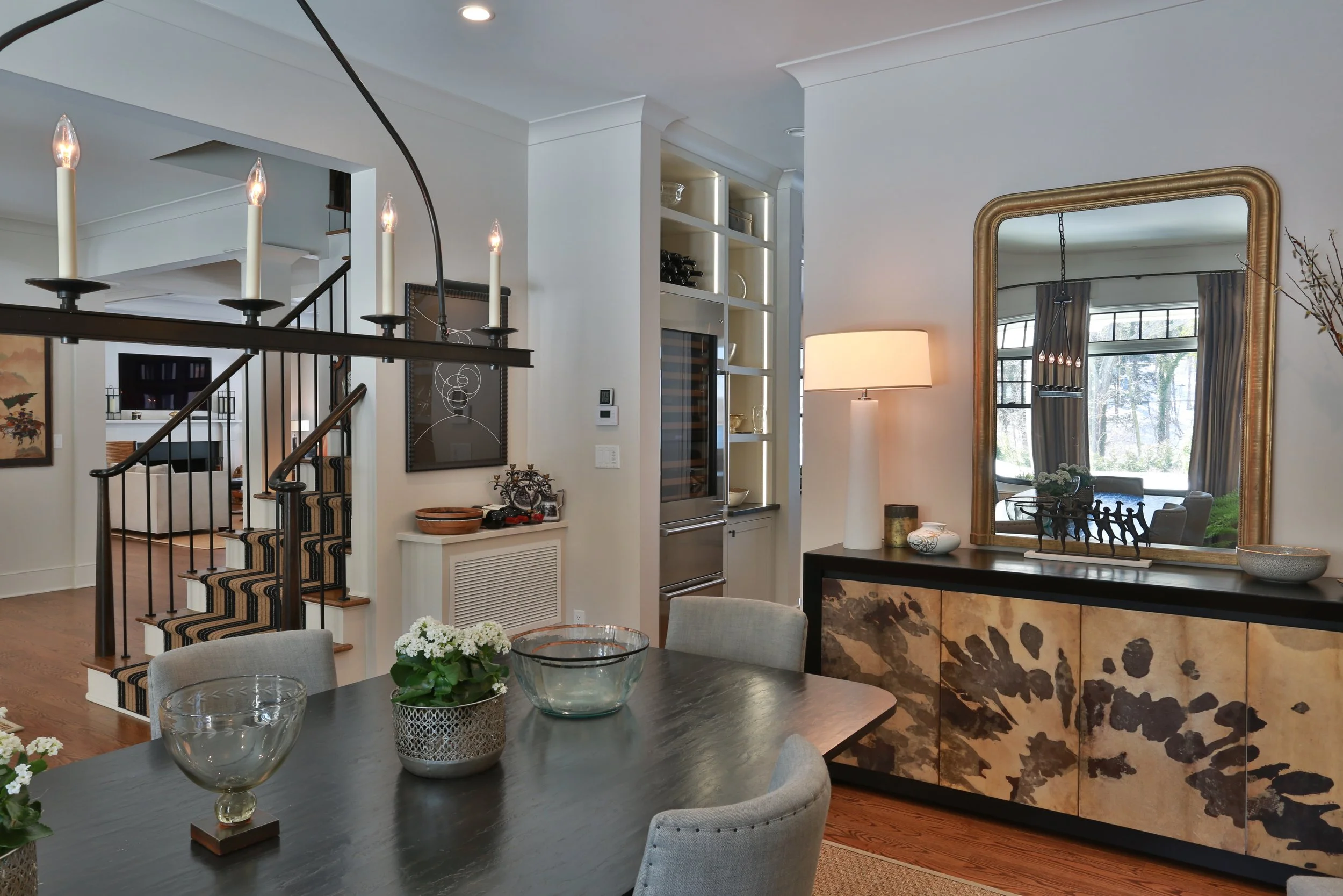 Interior of a modern dining and living area featuring a dark wood dining table with glass bowls, a large mirror with a gold frame, a black sideboard with decorative items, a floor lamp, built-in shelves with wine bottles, a staircase with striped car