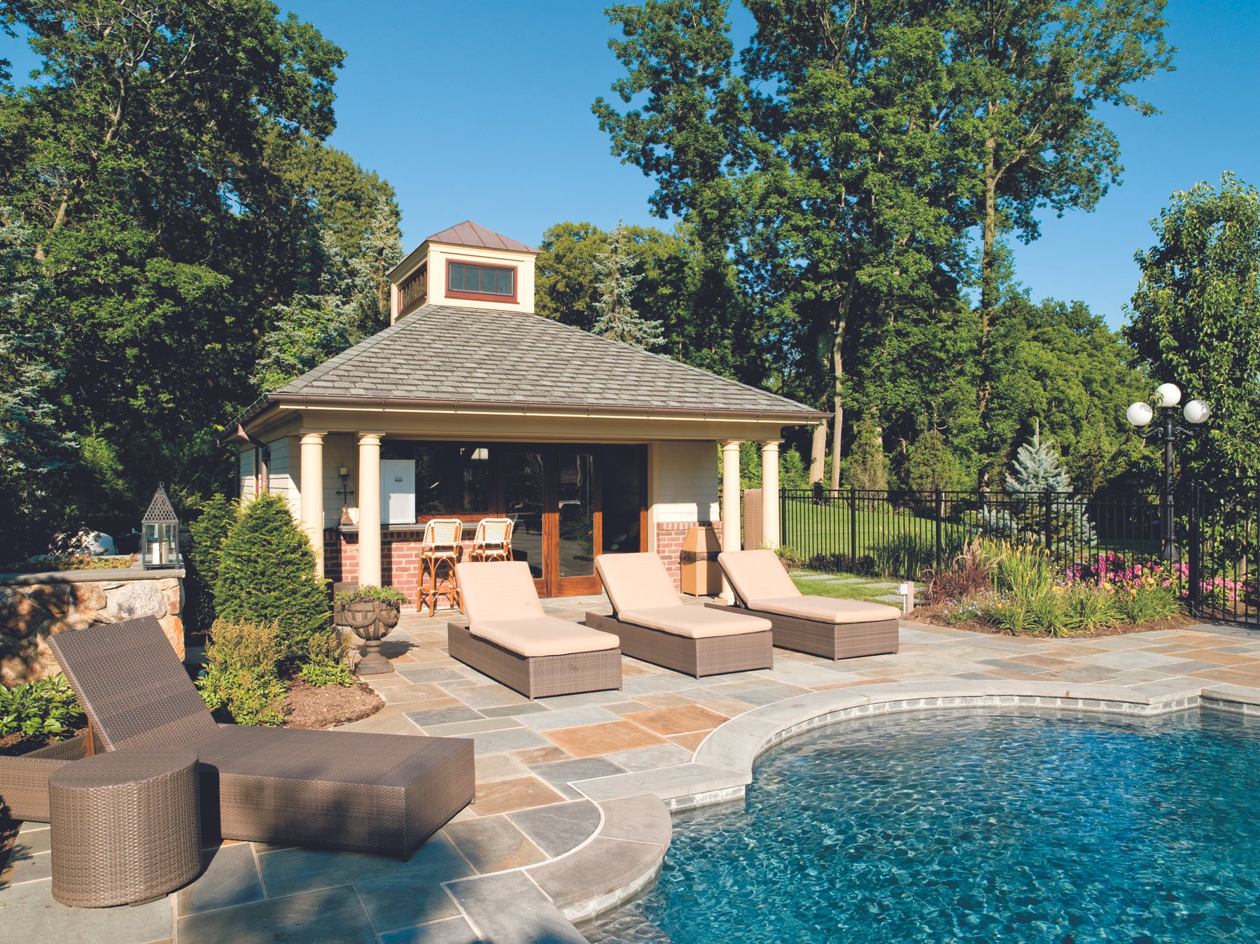 U-shaped swimming pool with beige lounge chairs and outdoor seating, a pool house with columns, surrounded by a landscaped yard with trees and plants.