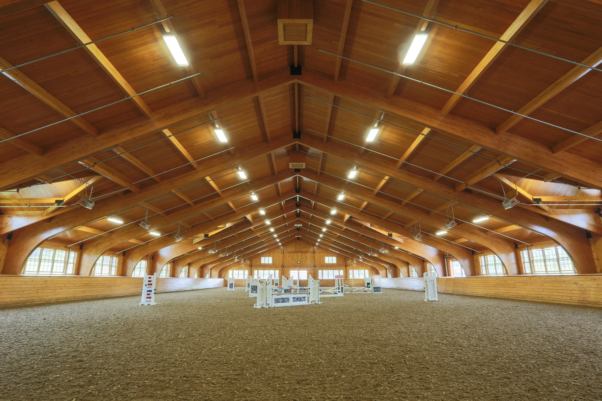 Indoor horse riding arena with a wooden ceiling and natural light from large windows, featuring equestrian jumps set up on the sandy ground.