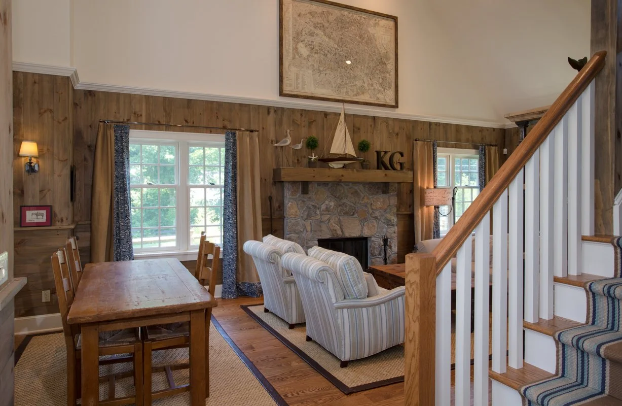 Living room with wood-paneled walls, a stone fireplace, striped armchairs, a dining table with chairs, and windows with curtains.