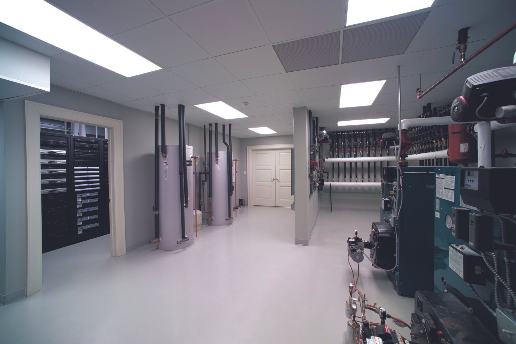 Industrial room with water heaters, electrical panels, and machinery, with a white door and ceiling lights.