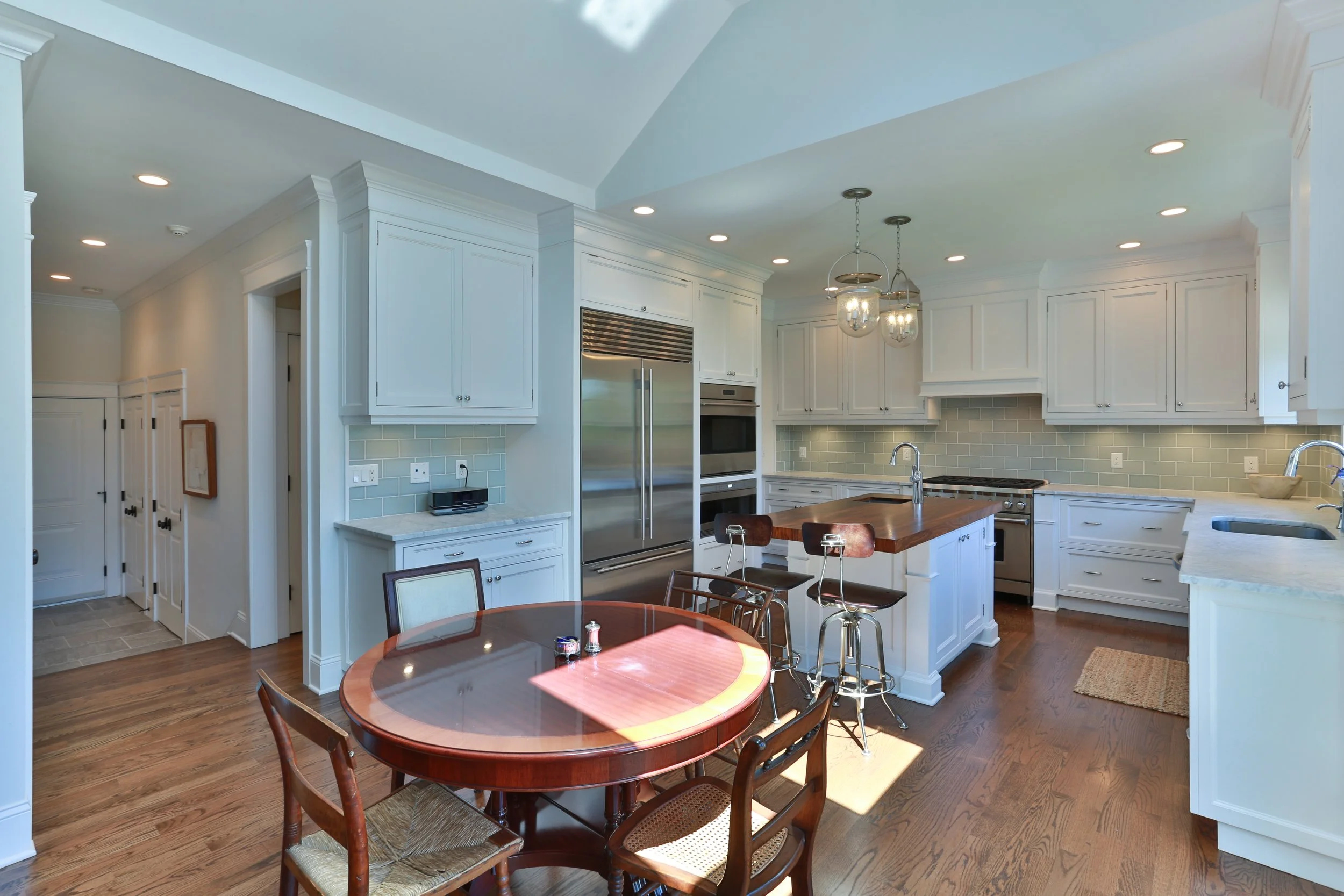 Bright kitchen with white cabinetry, wood flooring, and an island with a sink. There are two pendant lights hanging above the island, and a round wooden dining table with chairs. The kitchen features stainless steel appliances, including a refrigerat