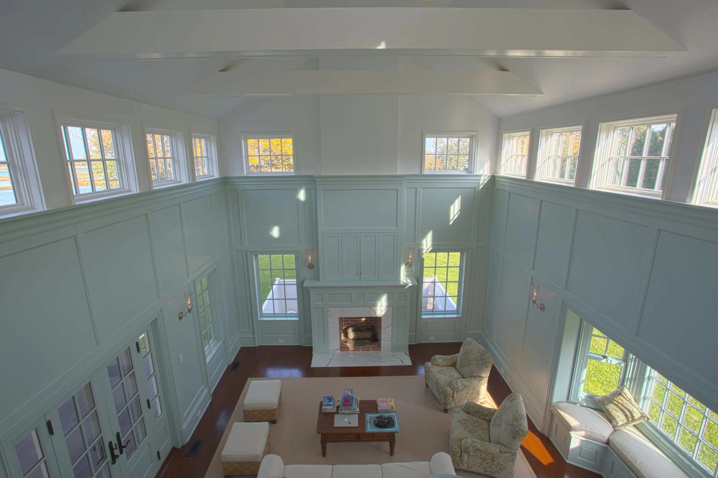 A spacious living room with high ceilings, large windows letting in natural light, a fireplace, and comfortable seating including an armchair and tufted ottomans.