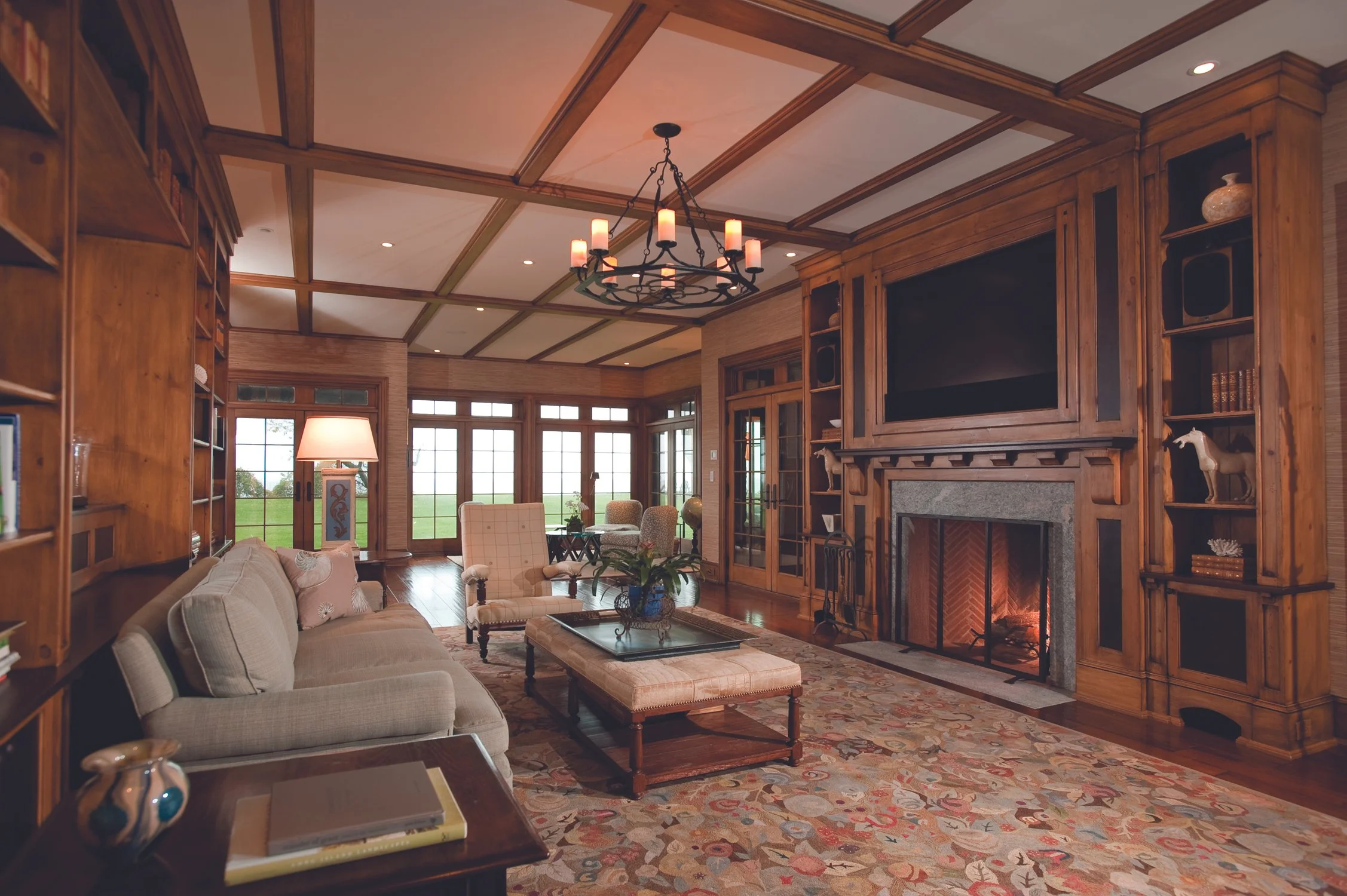 Living room with beige sofa, armchair, patterned area rug, wooden built-in shelves, fireplace, chandelier, and large windows showing green outdoor scenery.