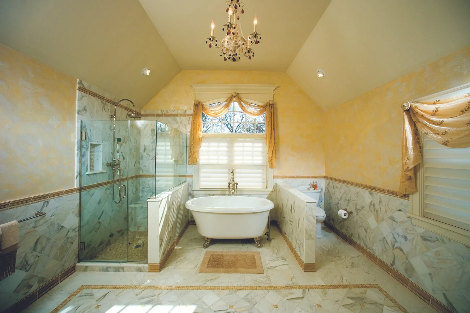 A luxurious bathroom with a freestanding bathtub, a glass-enclosed shower, marble floors, and ornate window treatments, with soft yellow and beige tones and a chandelier hanging from the ceiling.