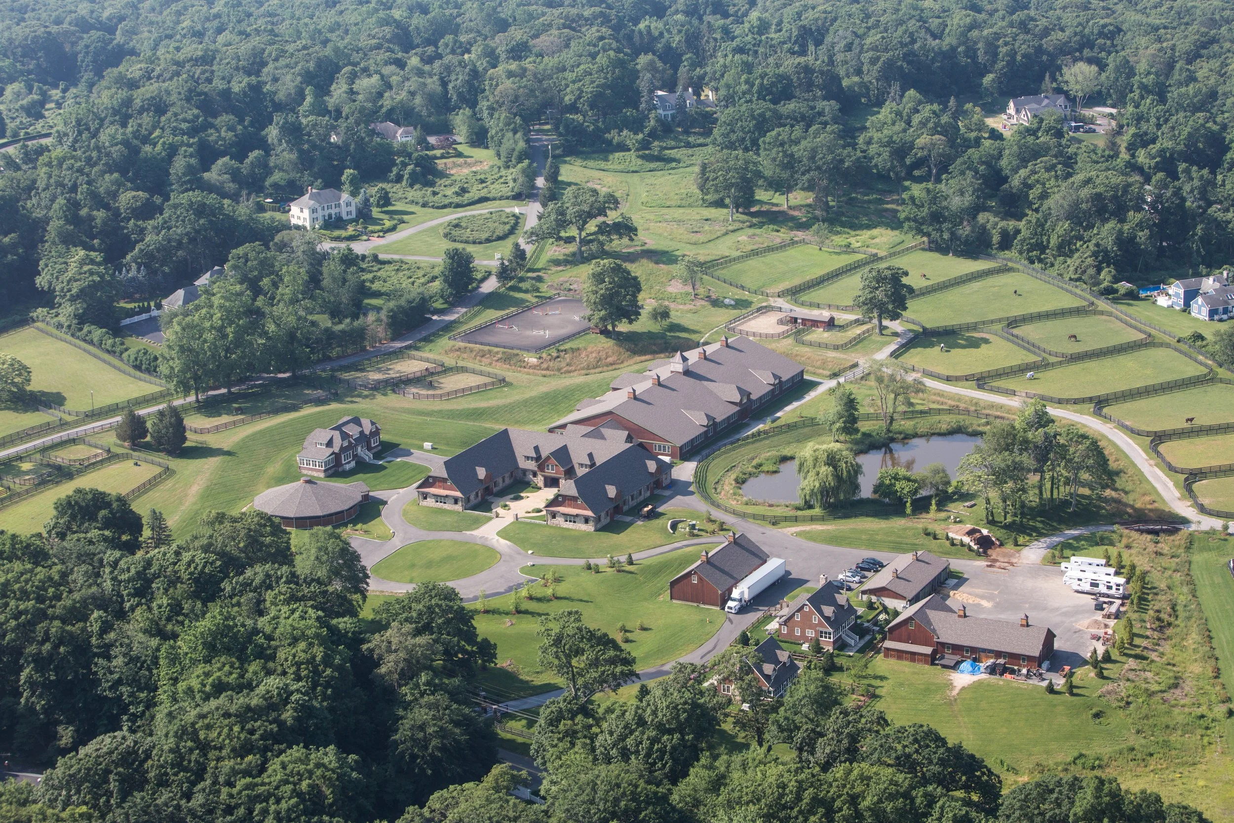 Aerial view of a large estate with multiple buildings, a pond, fenced horse pastures, and surrounding lush greenery.