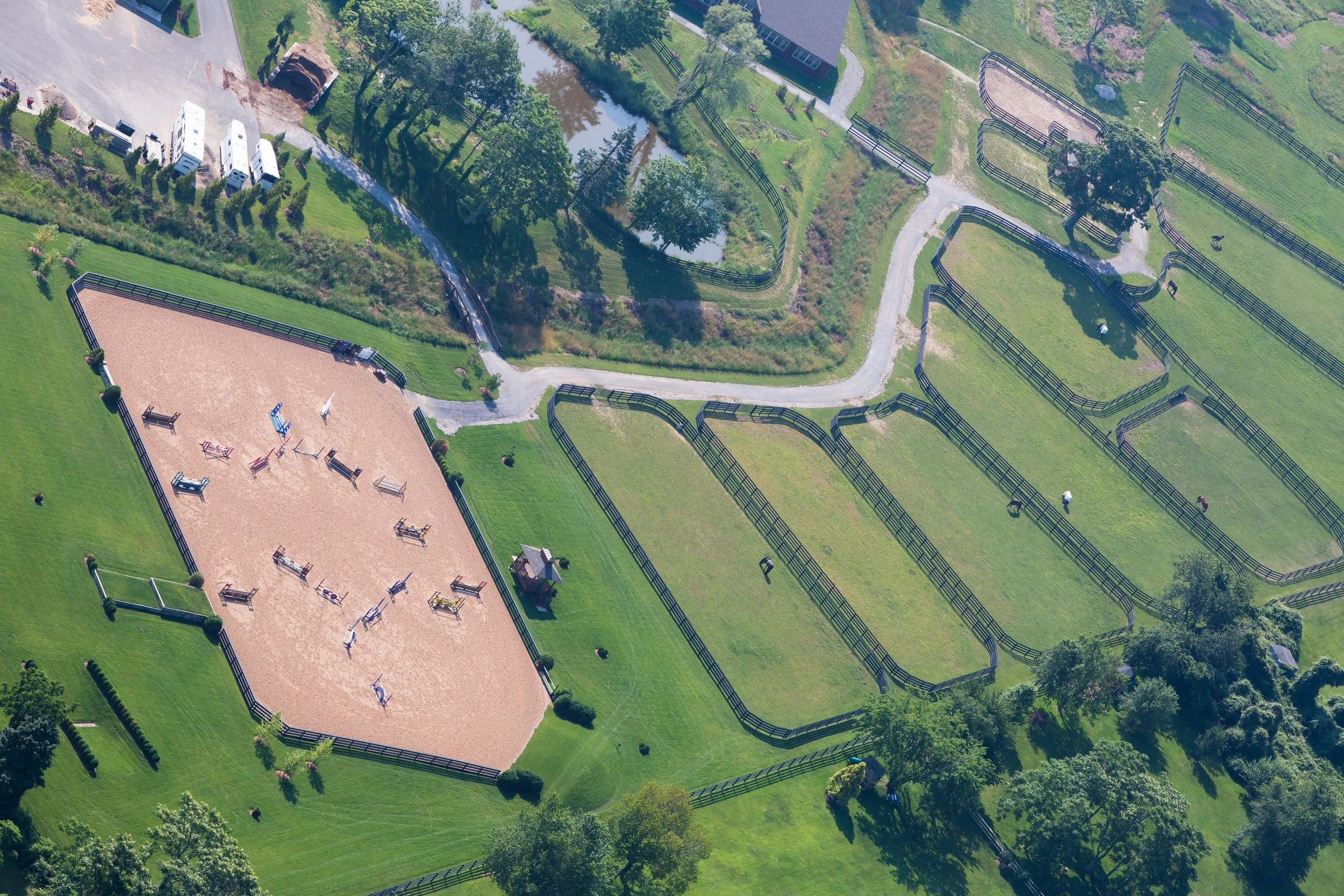Aerial view of a horse riding arena with jumps, surrounded by fenced grassy fields and trees, some with riders and horses grazing.