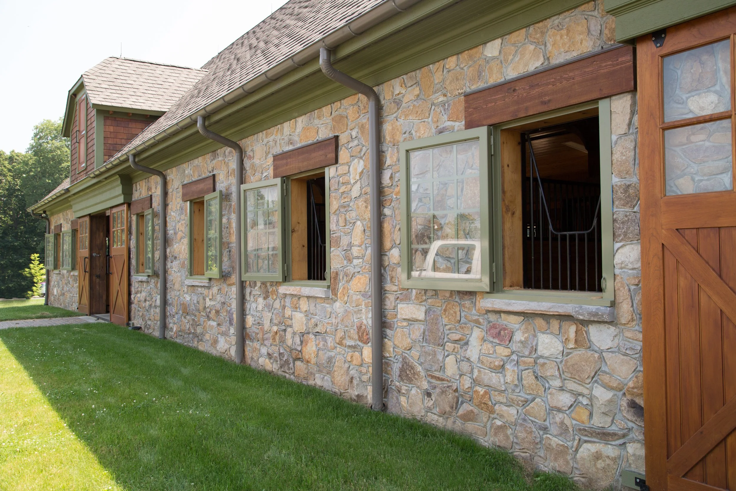Exterior of a stone building with wooden window shutters and a wooden door, green lawn in foreground, trees in background, and open windows showing black metal bars inside.