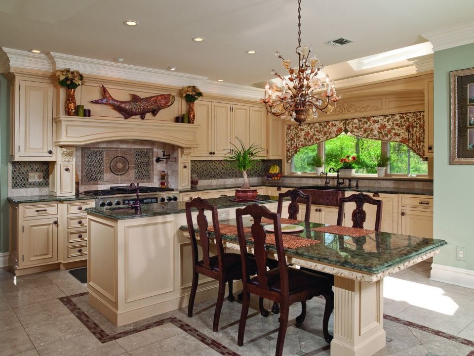 Kitchen with cream cabinets, granite countertops, a central island, and a dining table with four chairs. Decor includes a fish mounting above the stove, plants, and a chandelier.