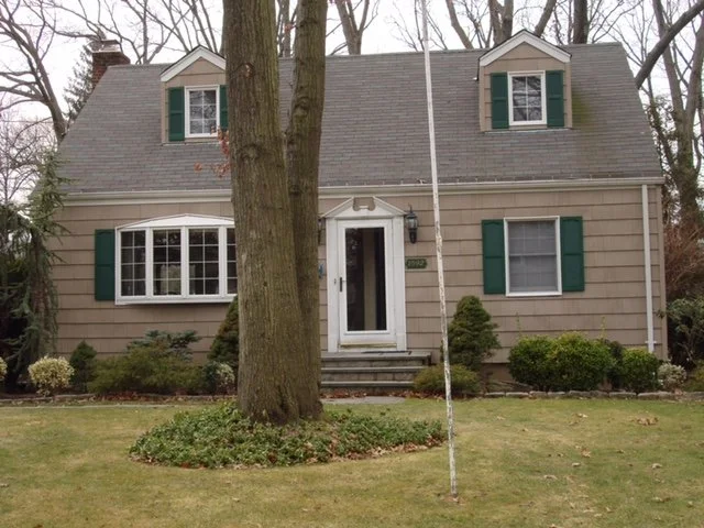 A beige two-story house with dark green shutters, a front door with steps leading up to it, a large tree in front, and a pole or flag in the yard.