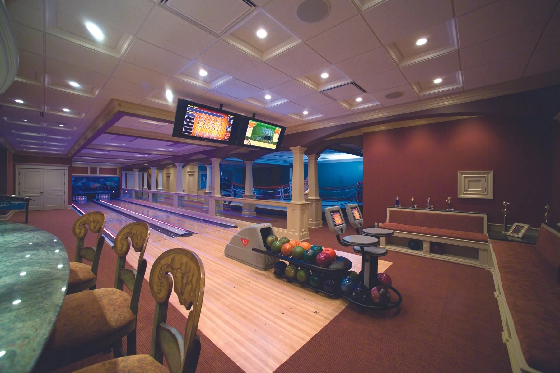 Indoor luxury bowling alley with wooden lanes, bowling balls in a rack, digital screens displaying scores, and seating area with chairs and a banquette.