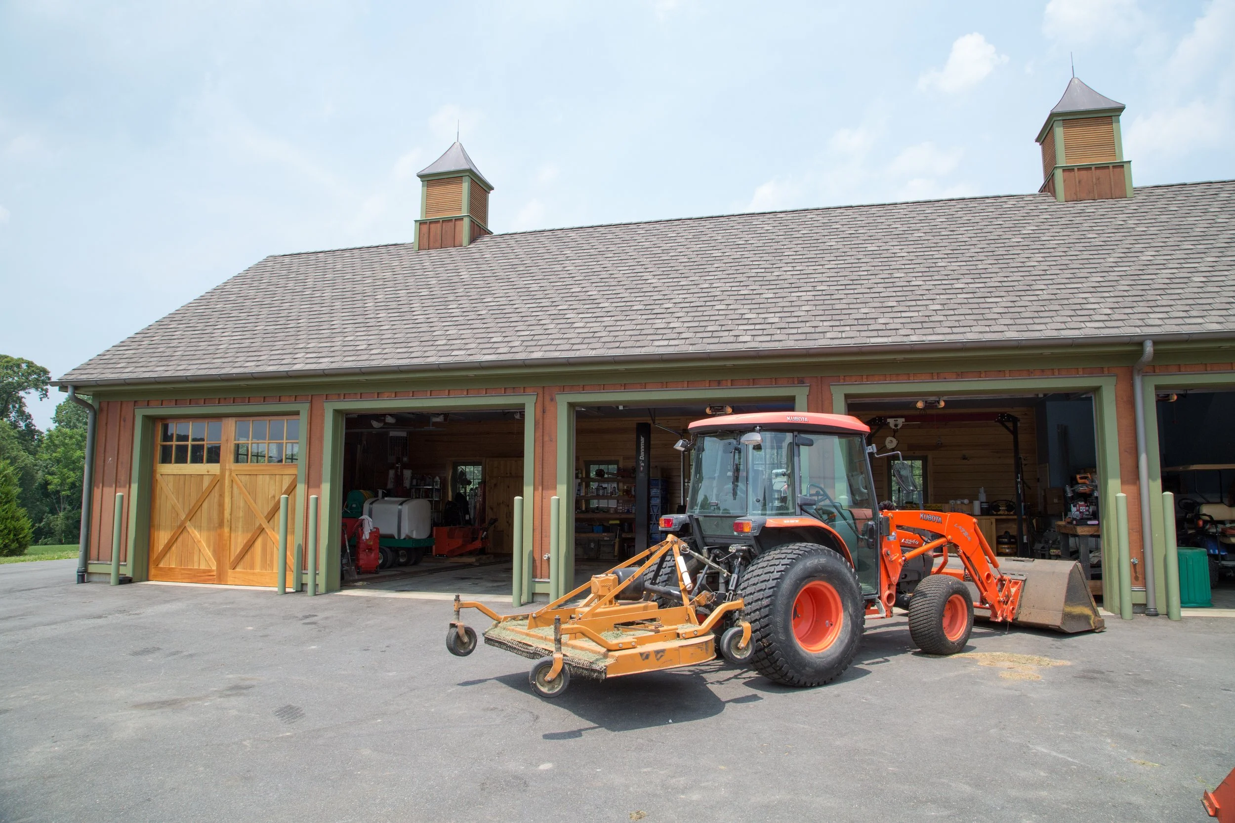 A garage with three open doors, a tractor with a mower attachment parked outside, and various tools inside, on a paved surface under a partly cloudy sky.