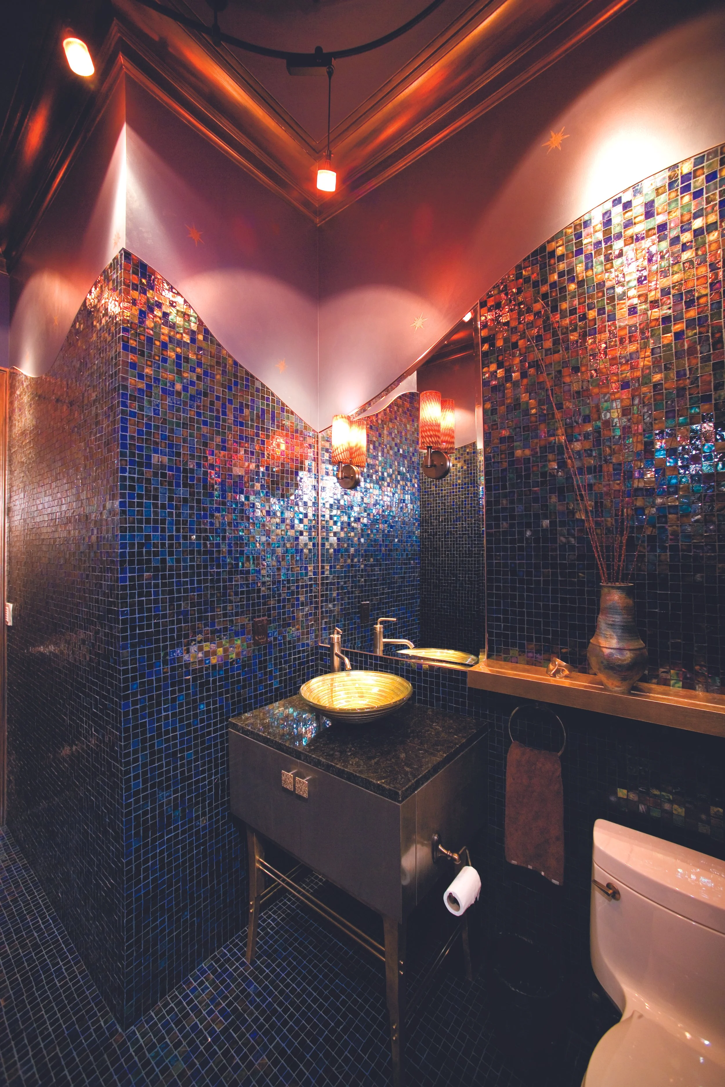 A bathroom with dark blue mosaic tile walls and ceiling, a gold circular sink on a black countertop, a mirror, a candle holder with lit candles, a vase with decorative branches, a hand towel, and a white toilet.