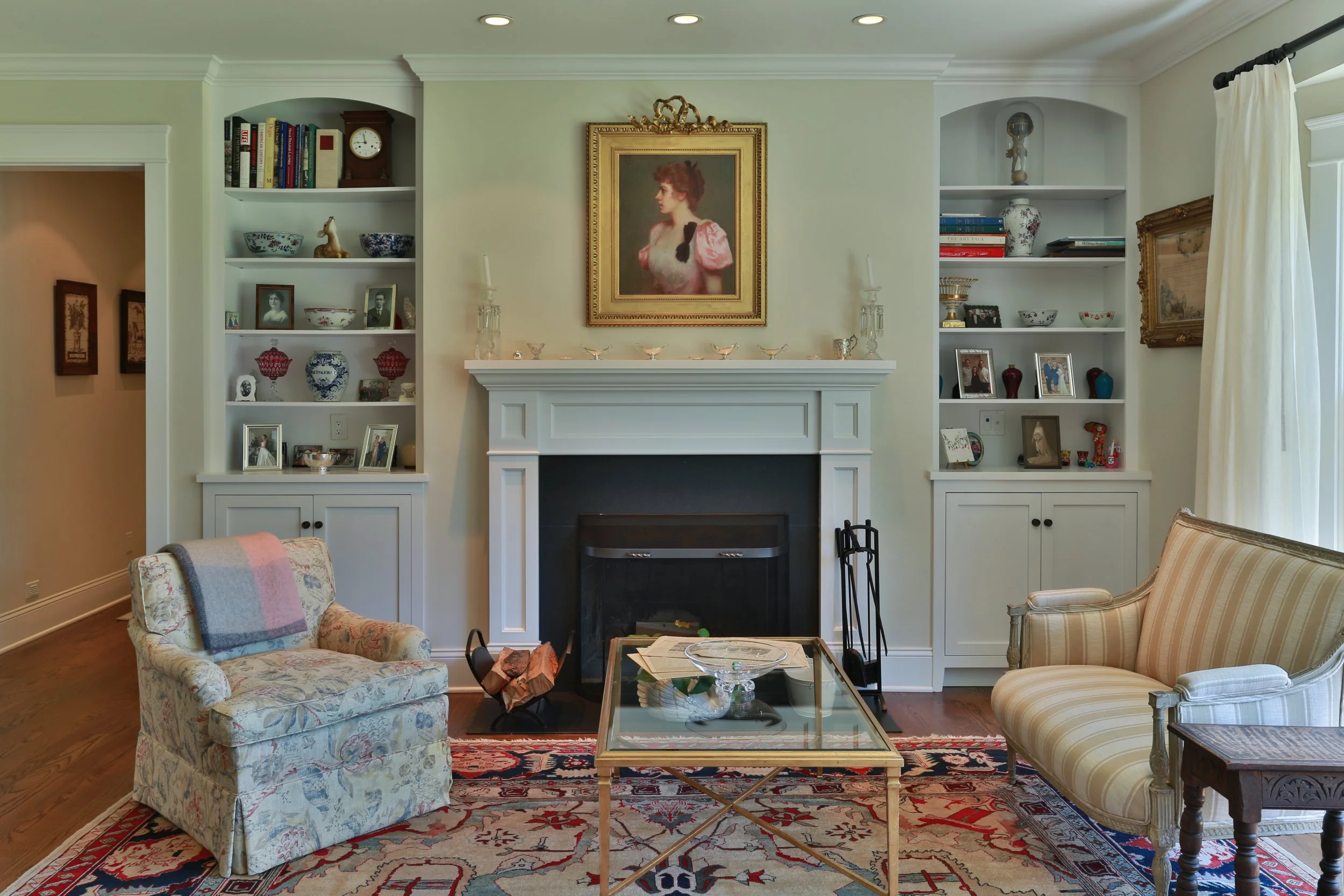 Living room with a fireplace, surrounded by built-in shelves filled with books, photographs, and decorative items. Two upholstered armchairs and a glass coffee table are positioned in front of the fireplace on a patterned rug. A large portrait of a w