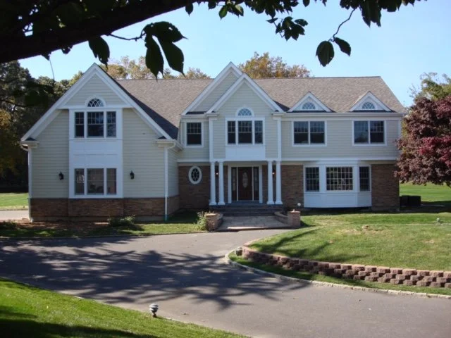 Large two-story house with white exterior, multiple windows, and a brick foundation, surrounded by a well-manicured lawn and trees.
