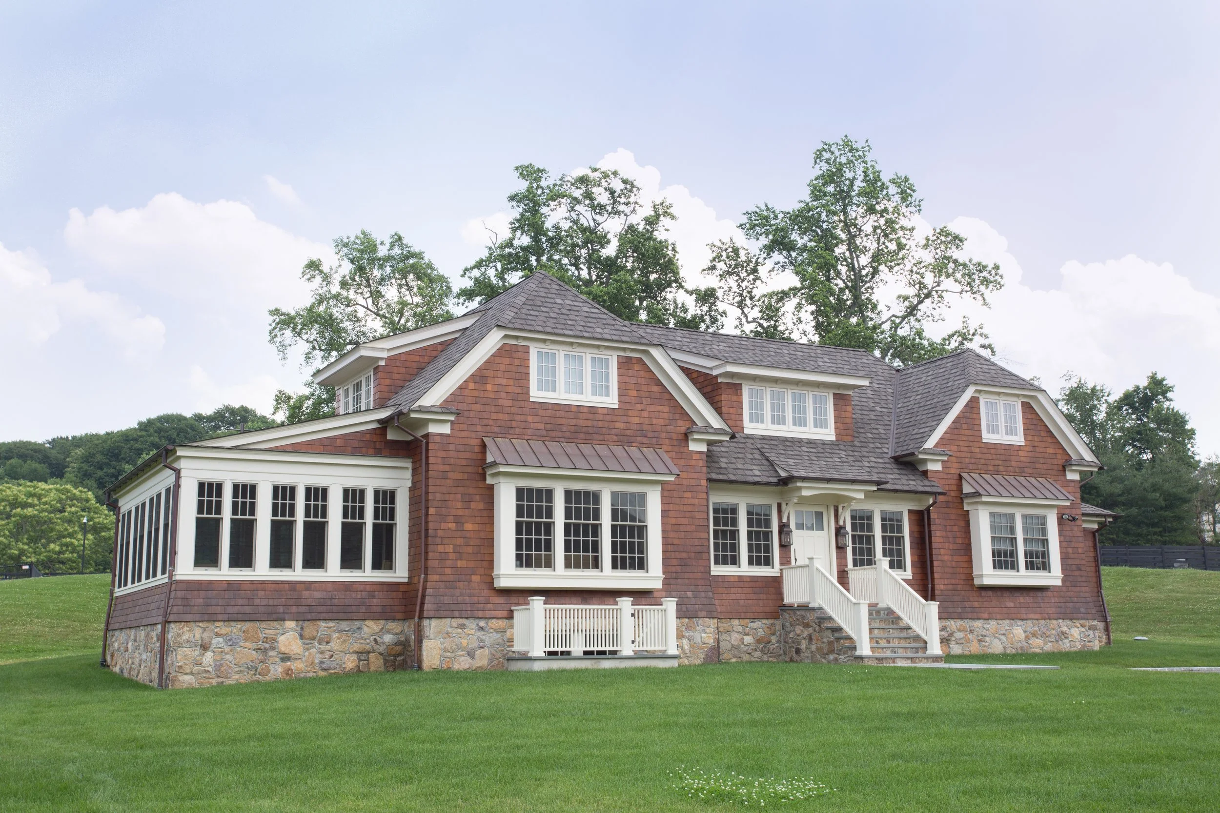 A large house with a stone foundation and a brown shingled exterior, featuring multiple windows and a front staircase with white railings, set on a well-maintained green lawn.