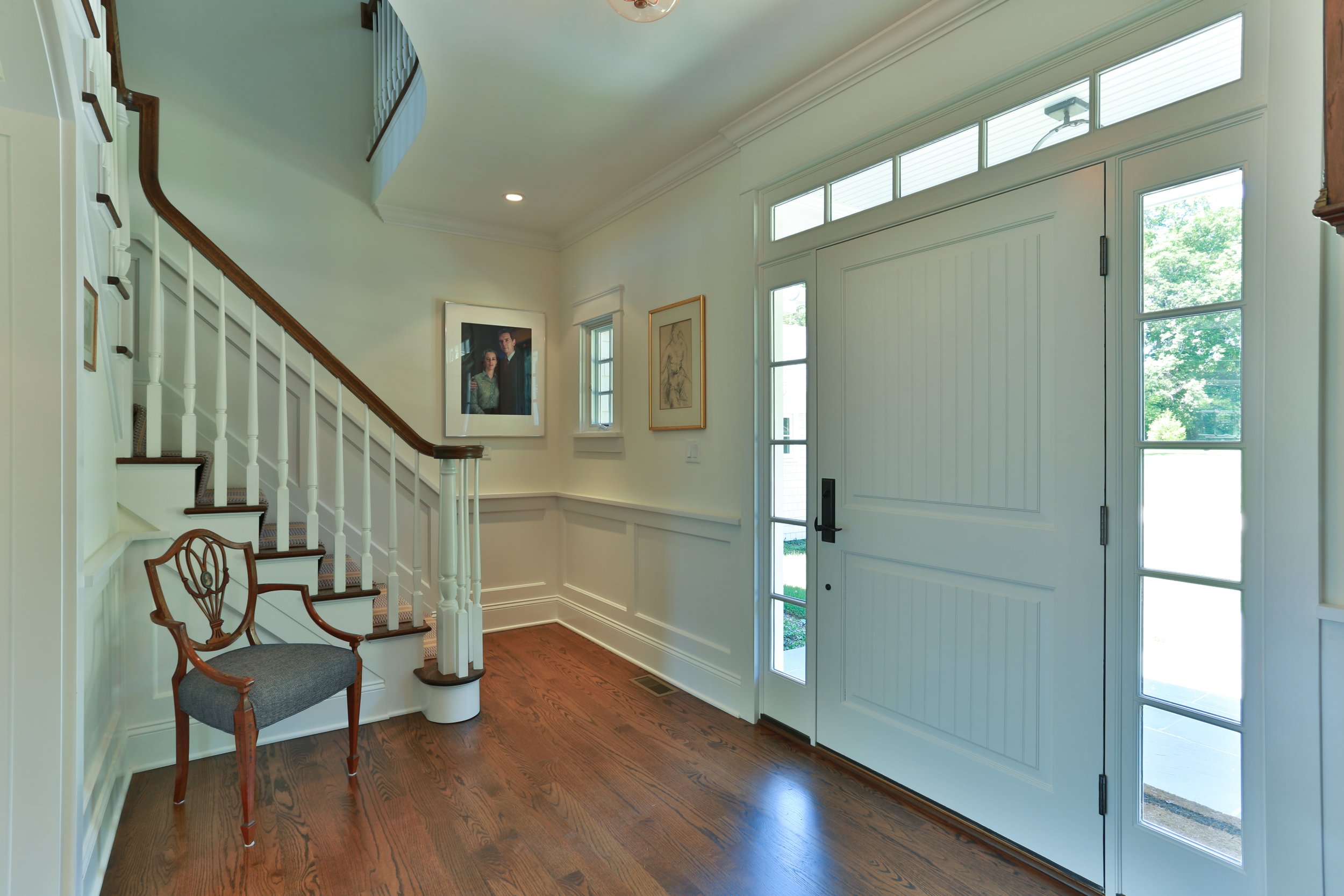 Entryway with a white front door surrounded by glass panels, hardwood floors, a staircase with white railing and wooden handrail, a vintage chair, and framed artwork on the walls.