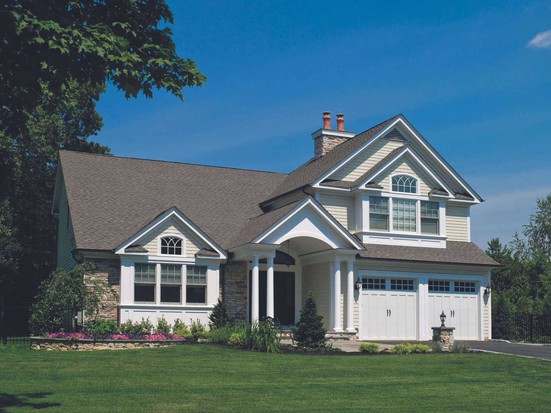 A two-story suburban house with beige siding, white trim, and a brown shingle roof. The house has a prominent front porch with white columns, a double garage with white doors, and landscaped front yard with green grass and flower beds.