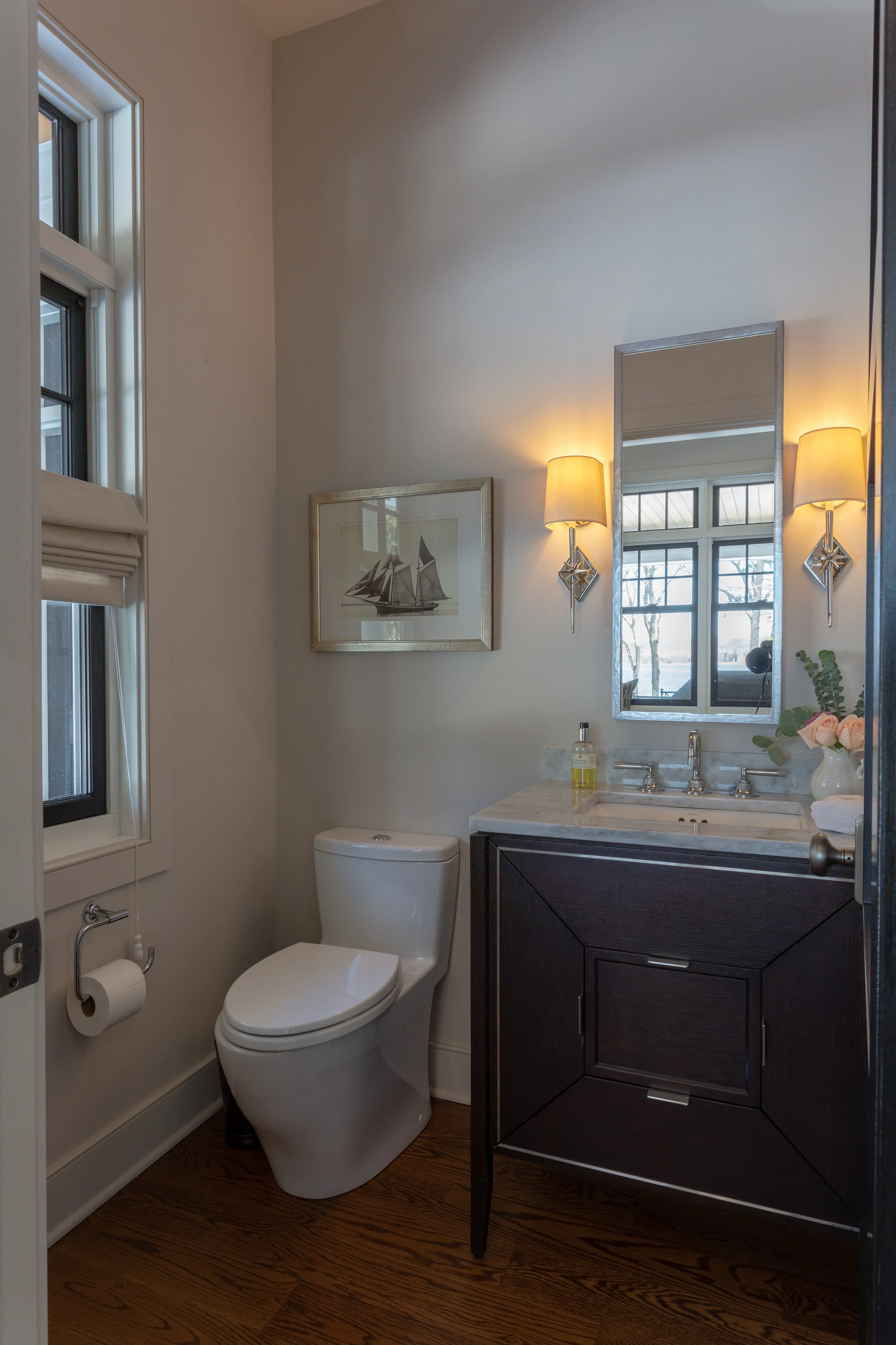 A modern bathroom featuring a toilet, a dark wood vanity with a marble countertop, a mirror, wall-mounted lights, a framed boat artwork, and a window with landscape view.
