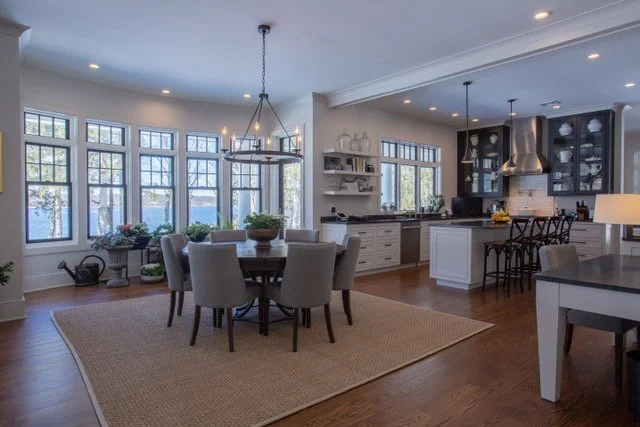 Open-concept kitchen and dining area with large windows, round dining table with six chairs, chandelier, white cabinets, black countertops, and bar stools.