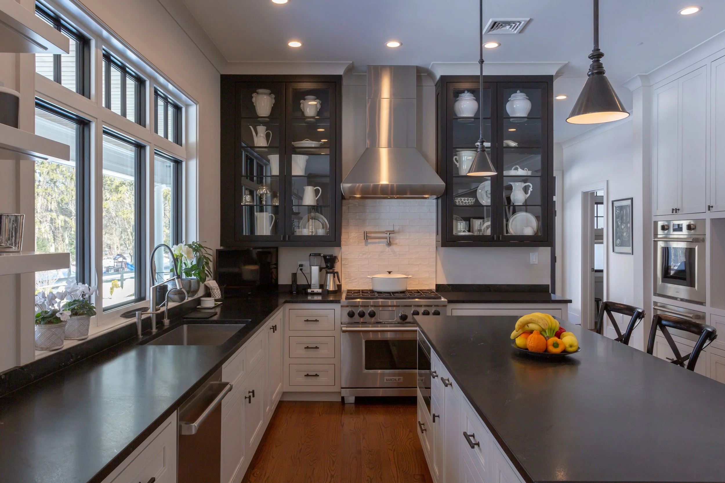 Modern kitchen with black countertops, white cabinets, stainless steel appliances, and a large window with a view outside. A bowl of assorted fruits sits on the island, and there are glass-front cabinets with white dishware.