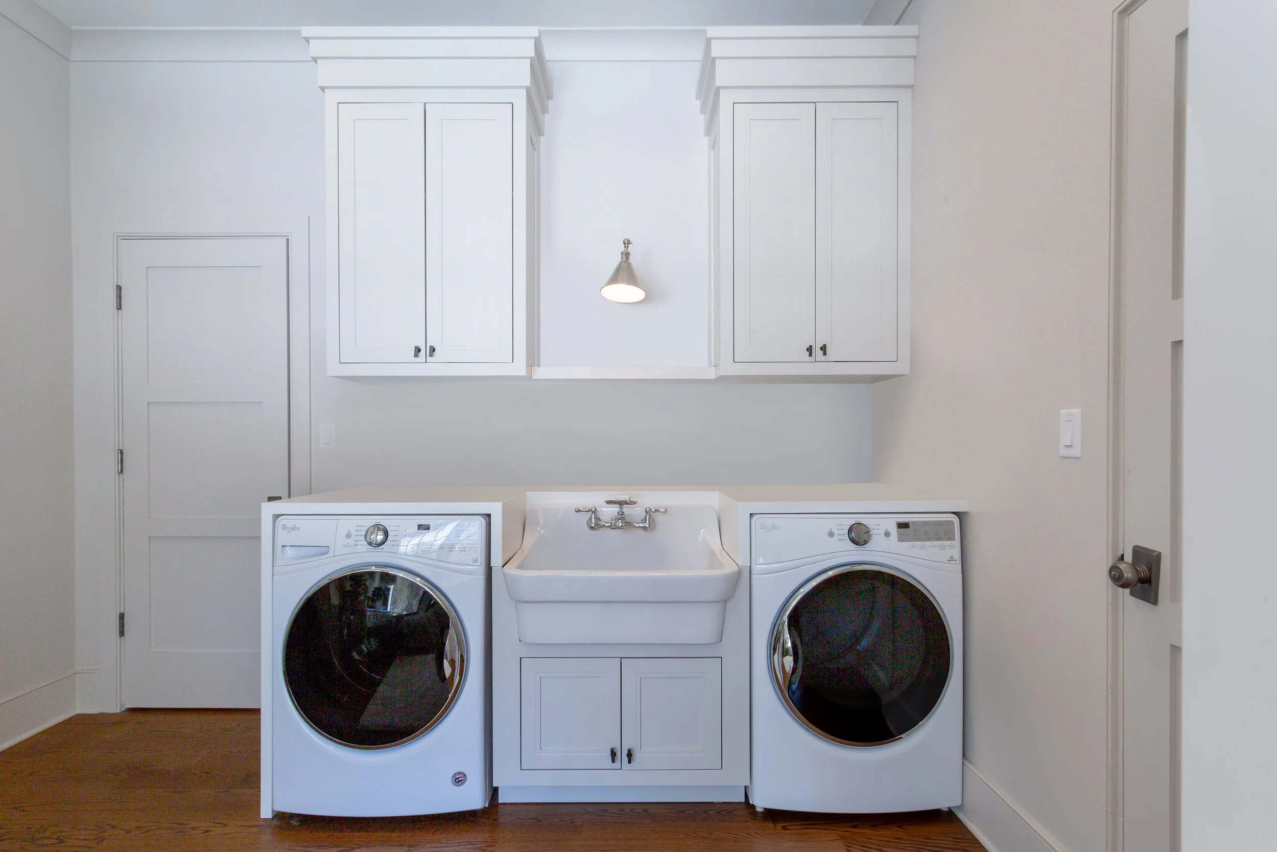 A laundry room with white cabinets, a utility sink, and a washer and dryer. The space has hardwood floors and a wall-mounted light fixture.