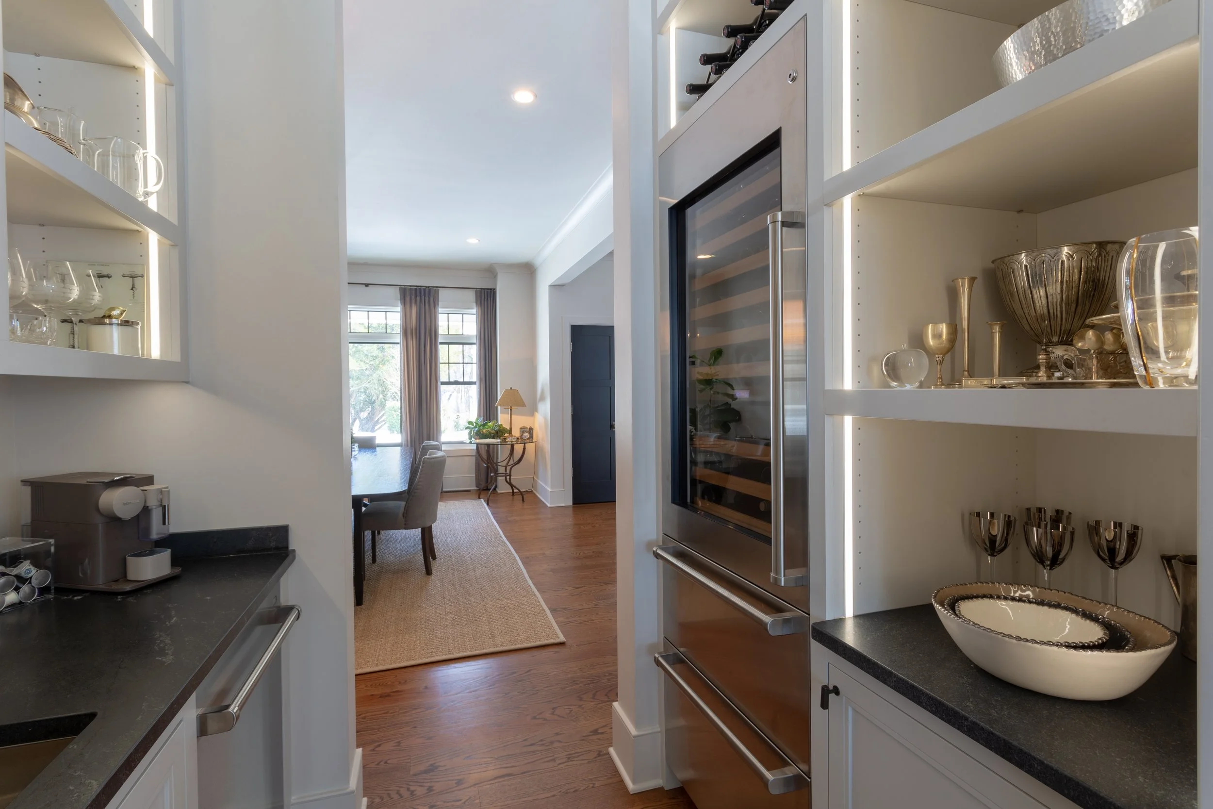 View from kitchen into dining and living area showing windows, curtains, table, chairs, rug, and side table with lamp.