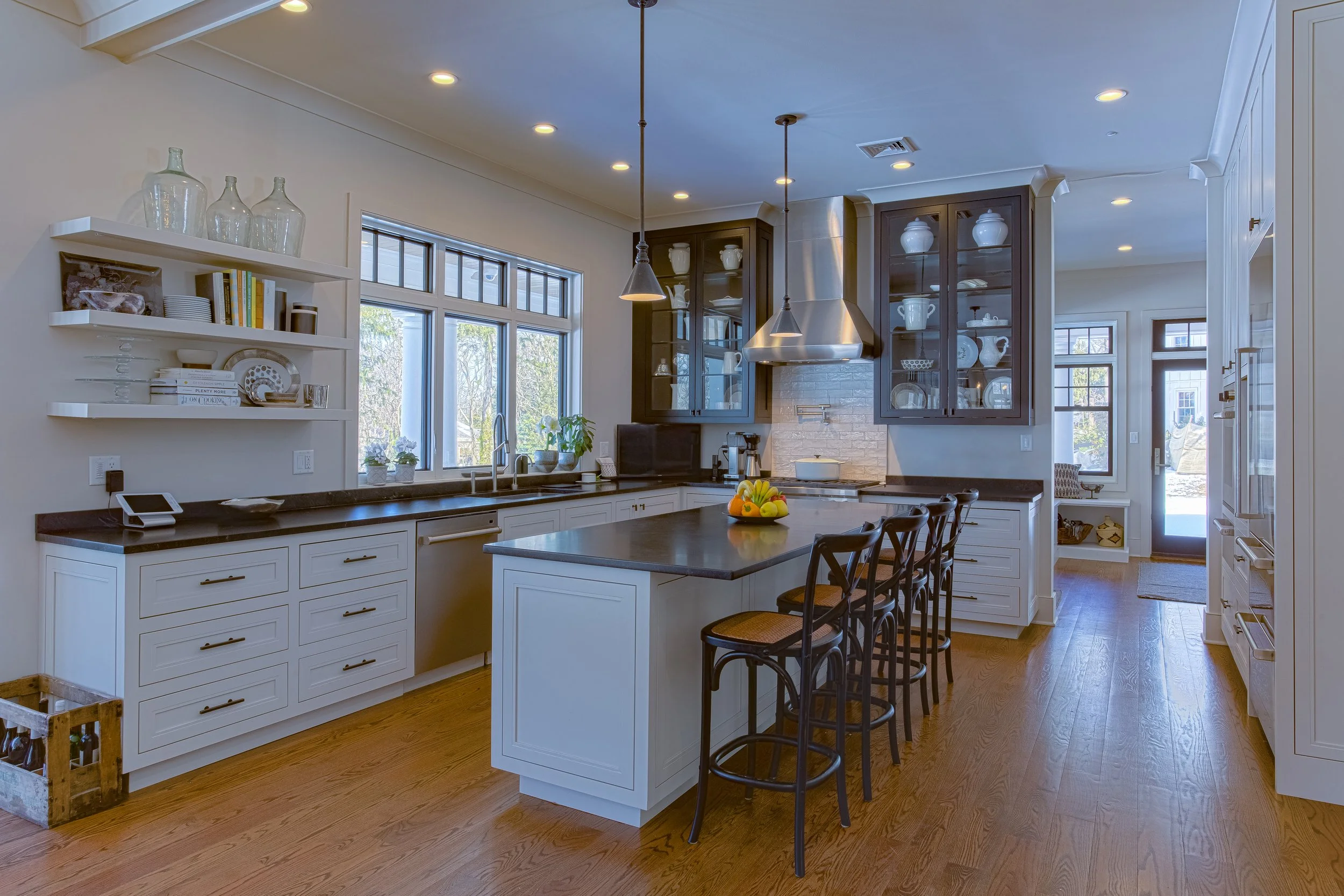 Modern kitchen with white cabinets, black countertops, a kitchen island, black chairs, java wood floors, and a large window, with open shelving and glass-front cabinets displaying dishware.