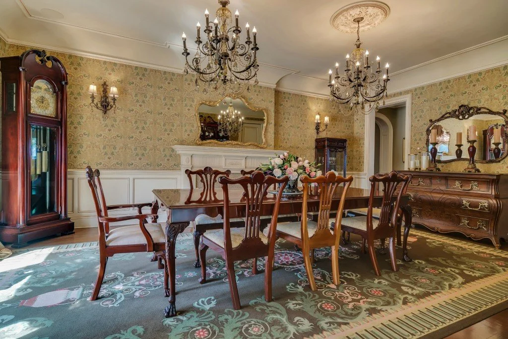 Elegant Victorian-style dining room with wooden furniture, floral wallpaper, chandeliers, a mirror, decorative vases, and a floral centerpiece on the table.