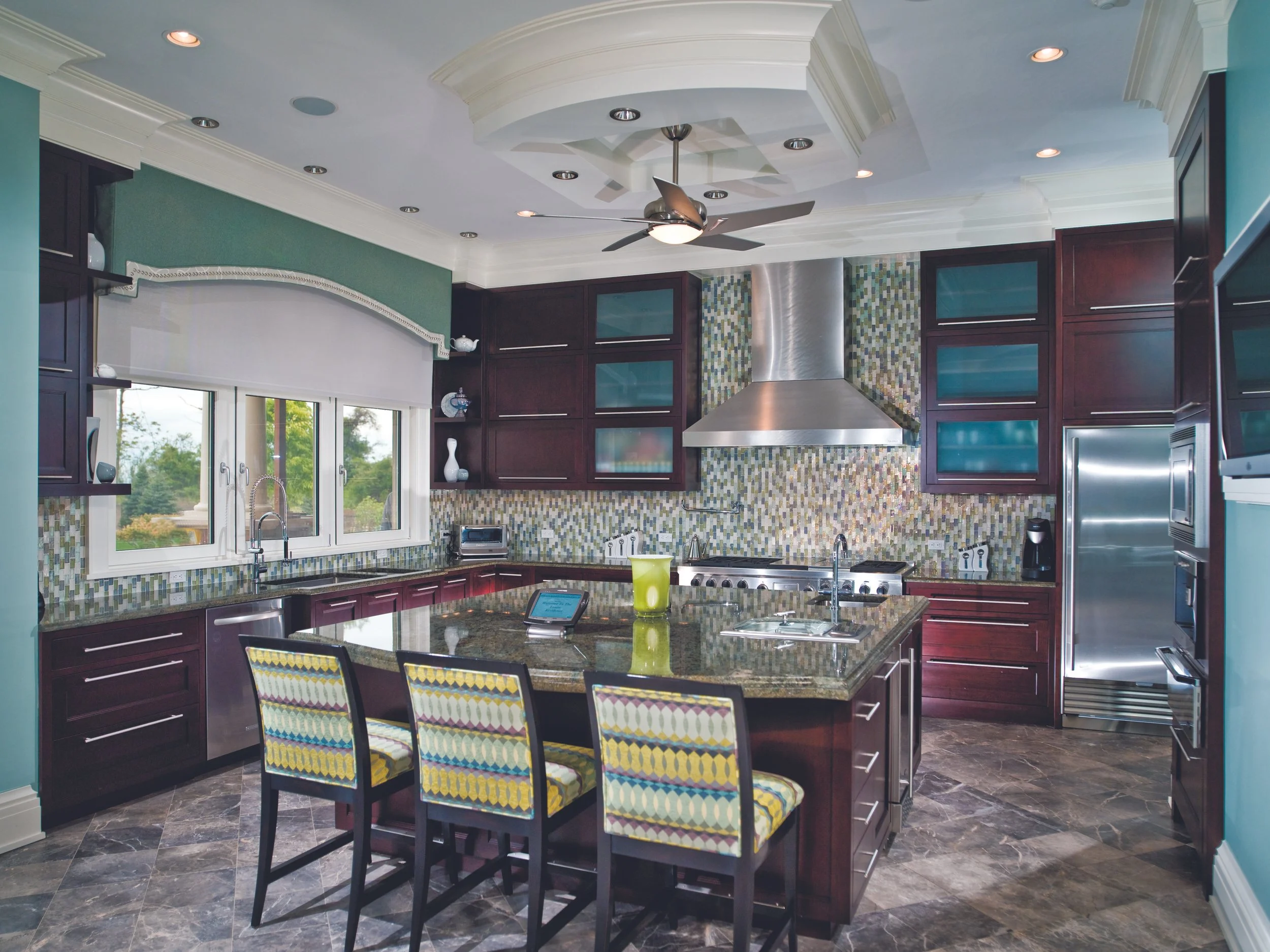 Modern kitchen with dark wood cabinets, a large granite island with four chairs, multicolored mosaic backsplash, stainless steel appliances, and a ceiling fan.