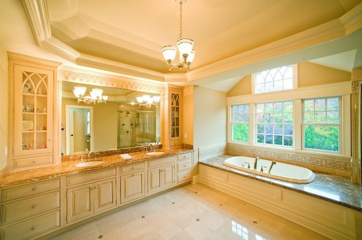 A spacious bathroom with a large built-in bathtub next to a window overlooking trees, white cabinetry, a marble countertop, and a large mirror, with soft yellow walls and a high ceiling with a chandelier.