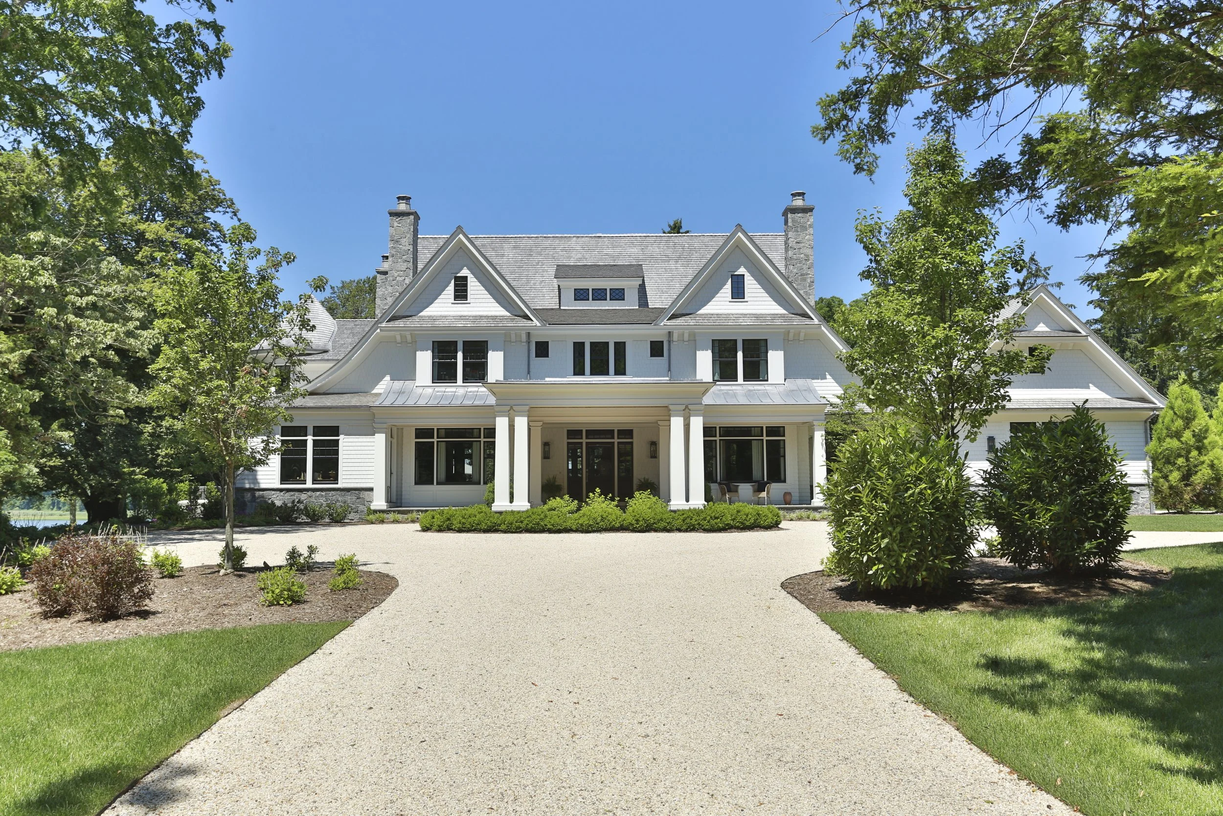 A large white house with multiple gables and chimneys, surrounded by trees and a landscaped yard, with a gravel driveway leading to the front entrance.