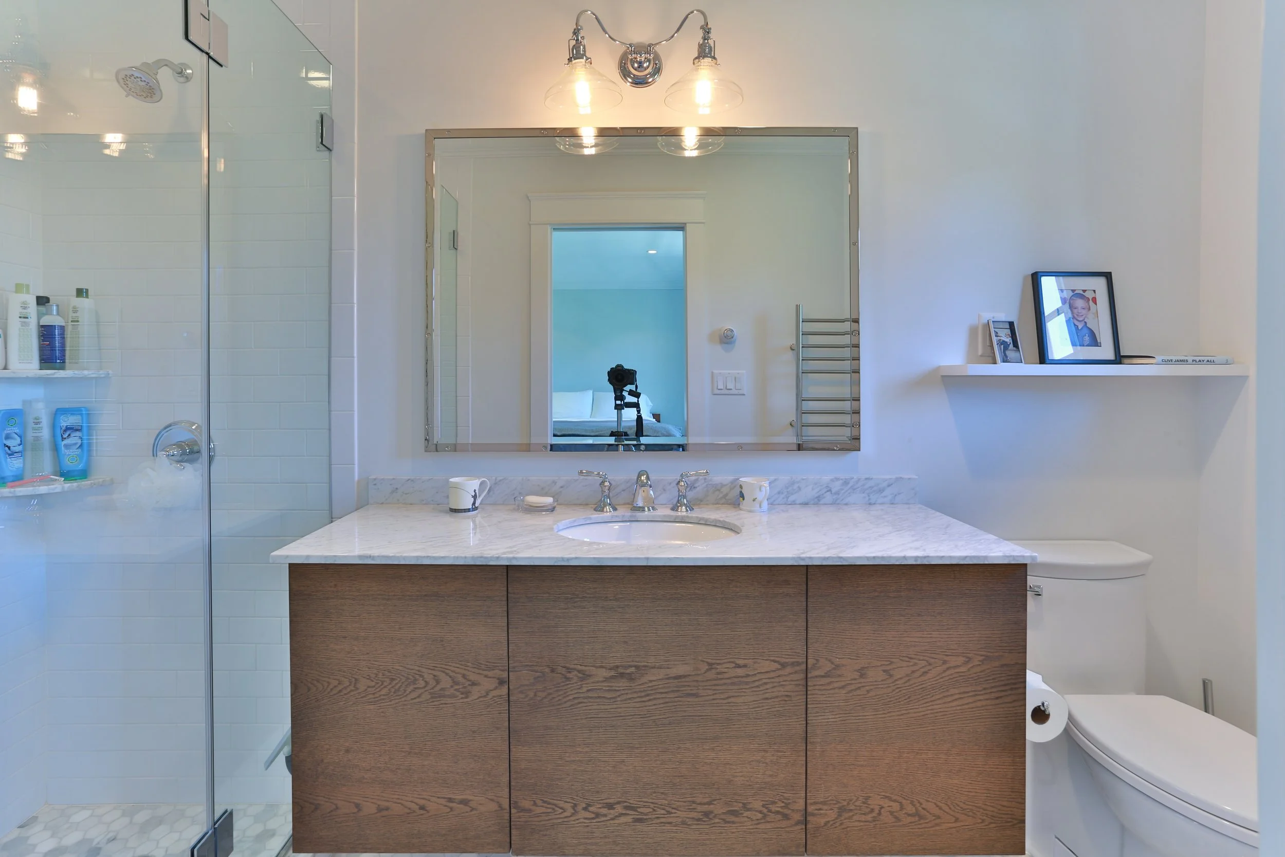 Modern bathroom with a marble countertop vanity, large mirror, wooden cabinet, shower with glass door, and a toilet, decorated with framed pictures on a white shelf.