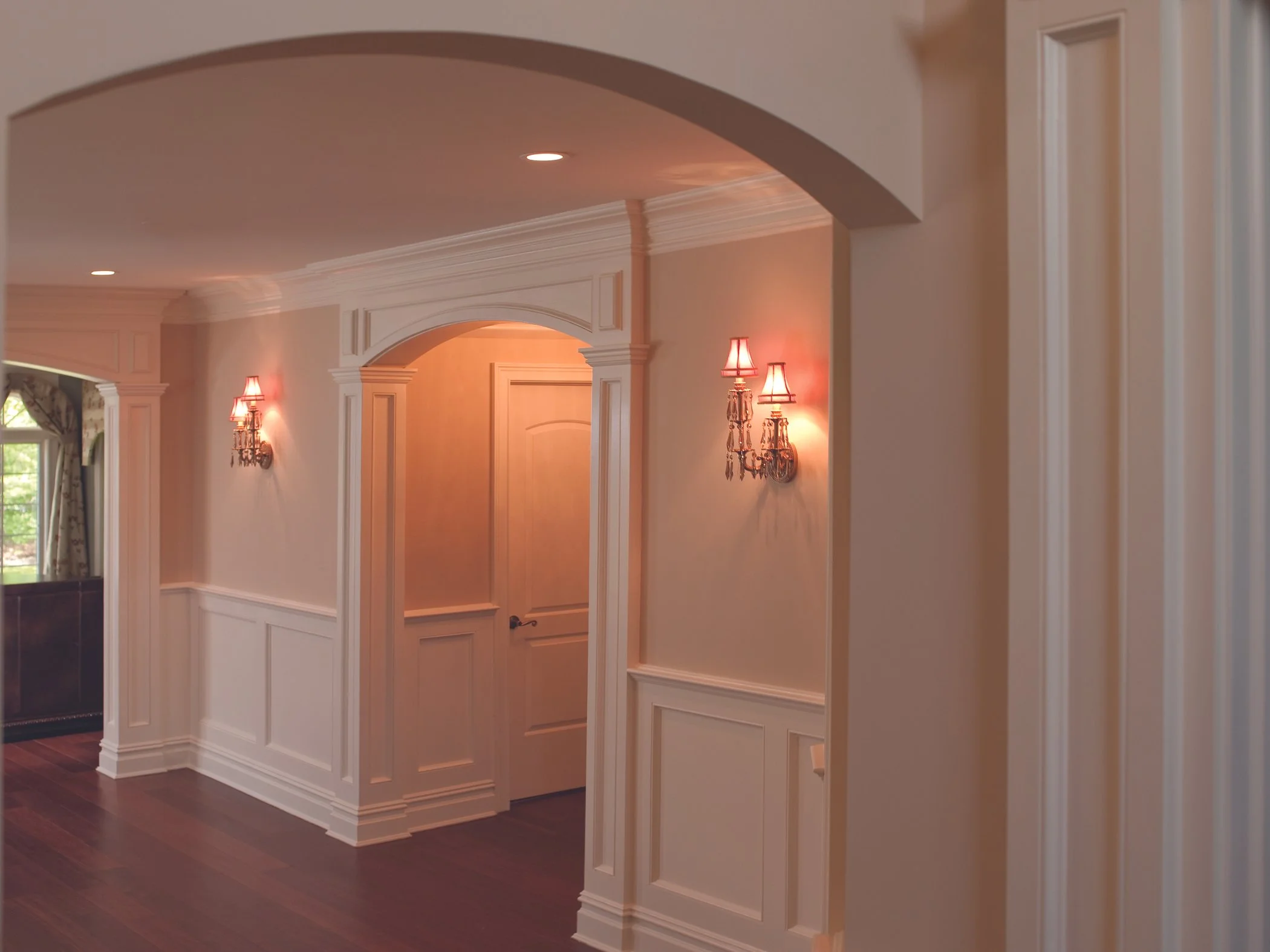 Interior view of a hallway with white paneled walls, decorative molding, two wall sconces with pink lampshades, and wood flooring.