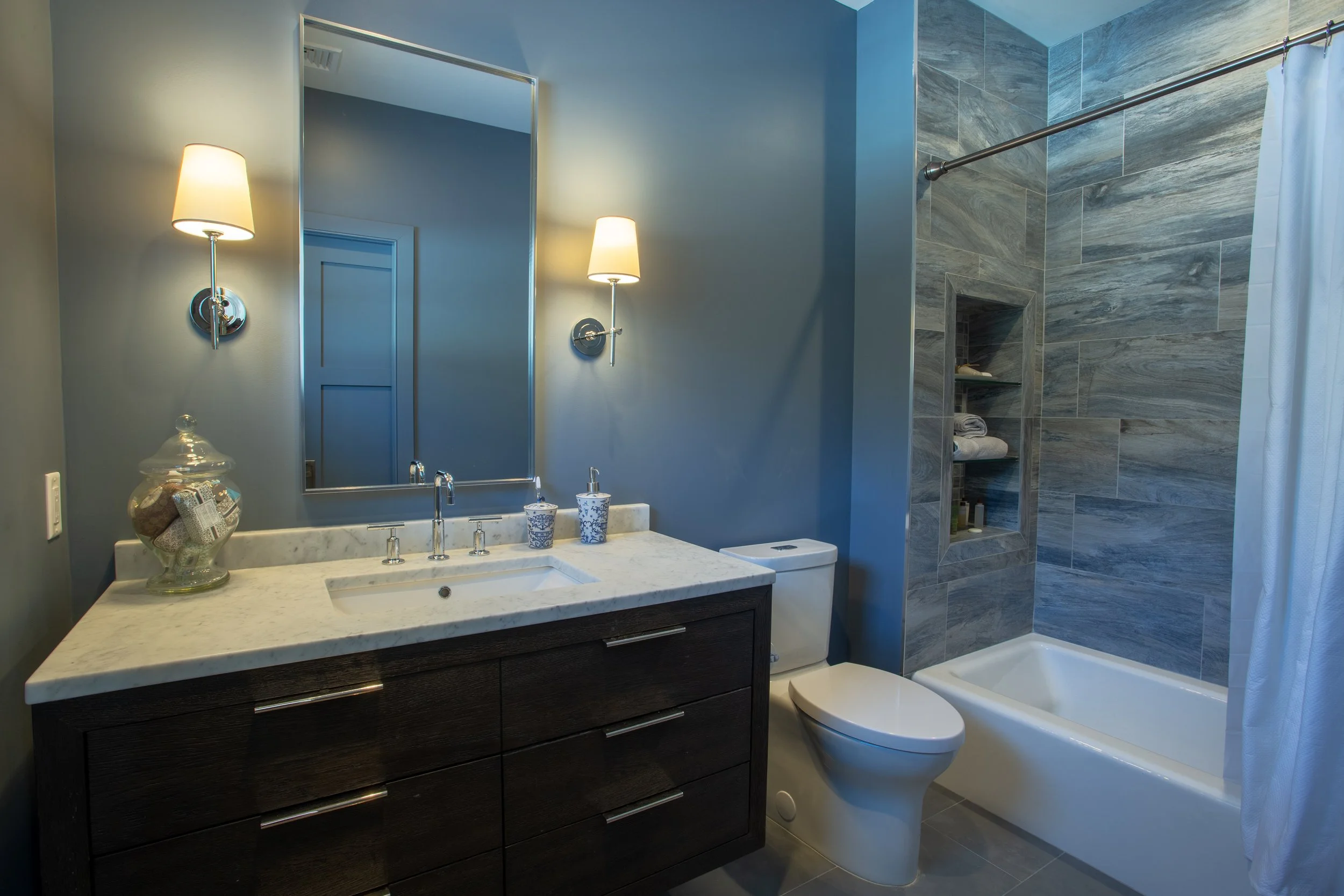 Modern bathroom with blue walls, marble countertop vanity, large mirror, two wall-mounted lamps, glass jar with toiletries, ceramic soap dispenser, white toilet, bathtub with gray tile surround, built-in shower niche, and a white shower curtain.