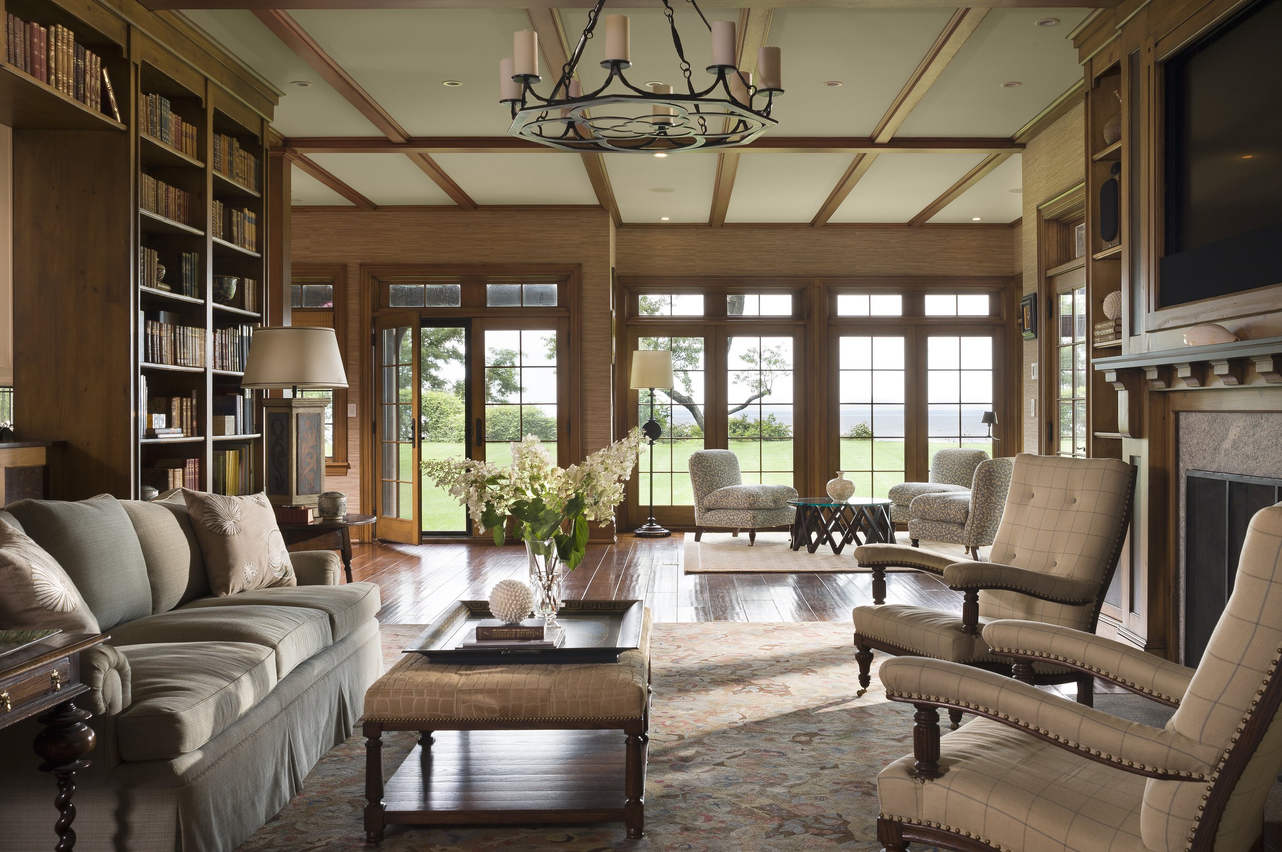 Bright living room with wooden floors, large windows, and comfortable seating including a sofa and armchairs, decorated with a chandelier, lamp, and plants.