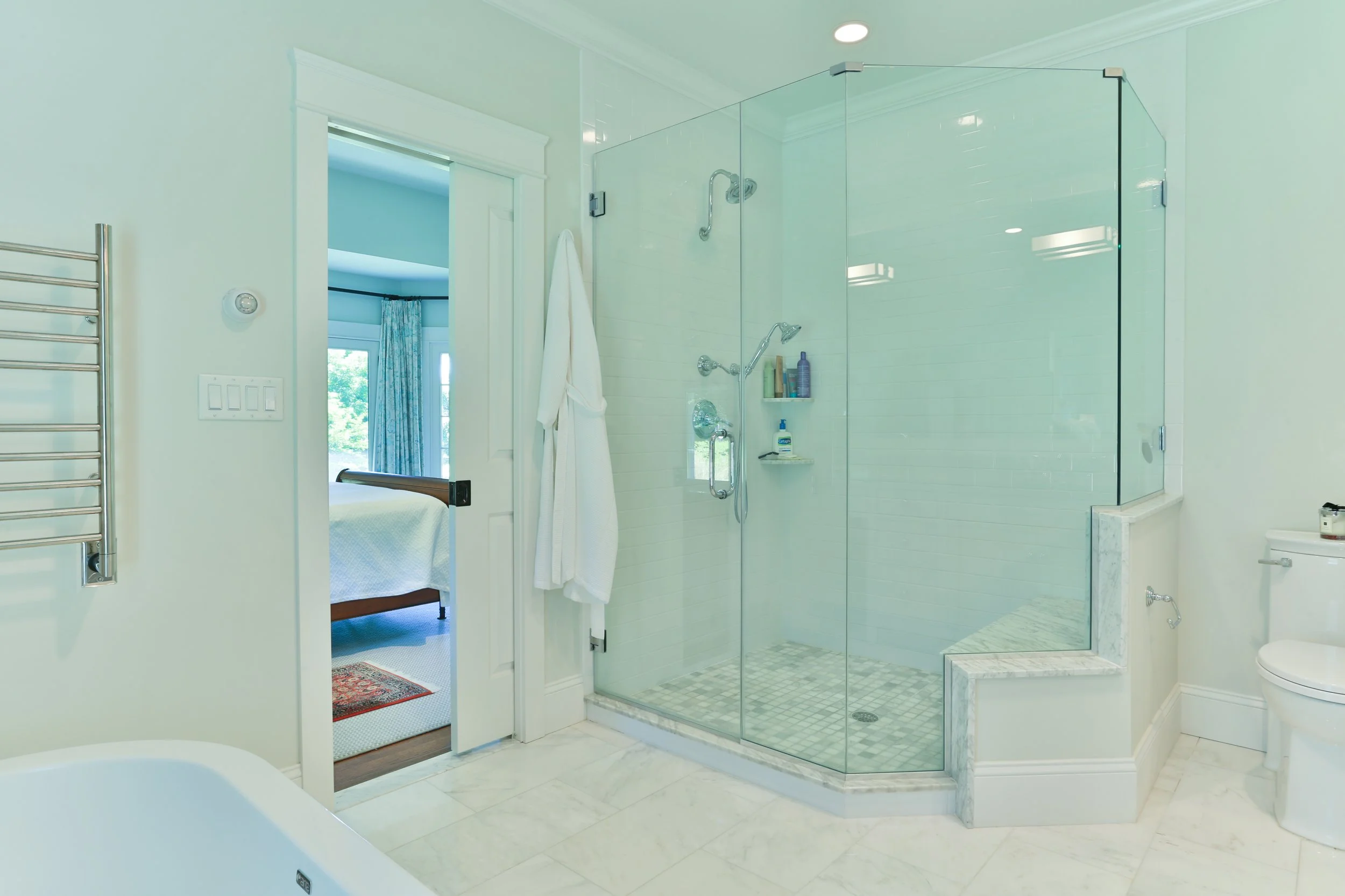 Bathroom with a glass shower enclosure, a white toilet, a towel rack, and an open door leading to a bedroom. The room has light-colored walls and marble flooring.