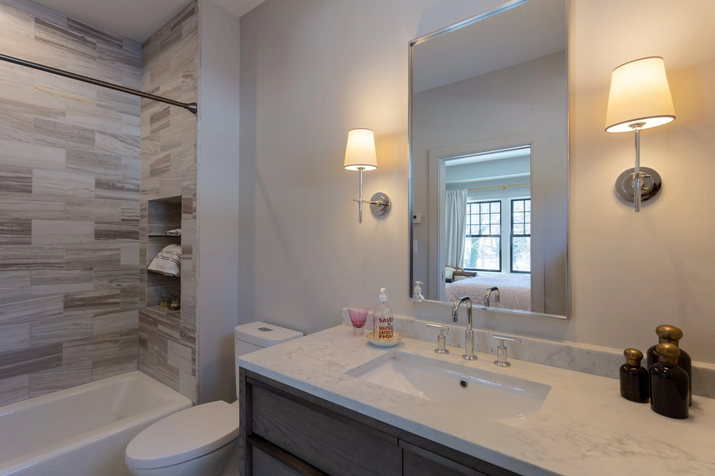 Modern bathroom with marble countertop, mirror, wall-mounted lamps, toilet, and partial view of a bedroom through the mirror reflection.