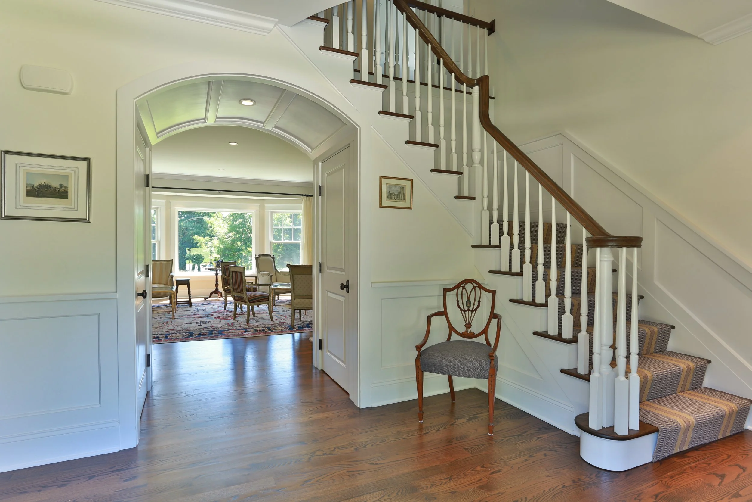 Interior view of a home showing a staircase with white railings and dark wood steps, an arched doorway leading to a sunlit dining area with large windows and a view of greenery outside, a wooden chair with a gray cushion, and framed pictures on the w