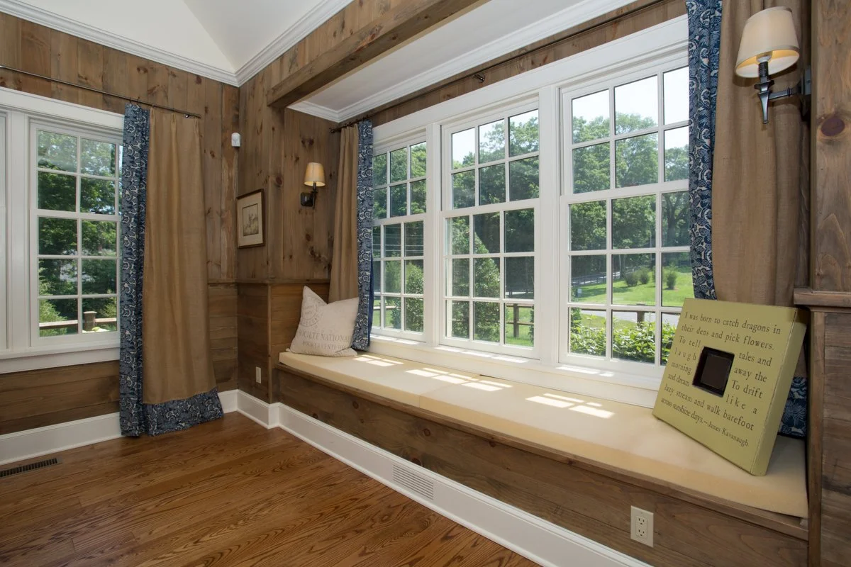 A cozy window nook with a cushioned seat, a decorative pillow, and a piece of artwork on the wood-paneled wall. Large windows reveal green trees outside, with plaid and beige curtains flanking the windows. A small light fixture on the wall and a poem