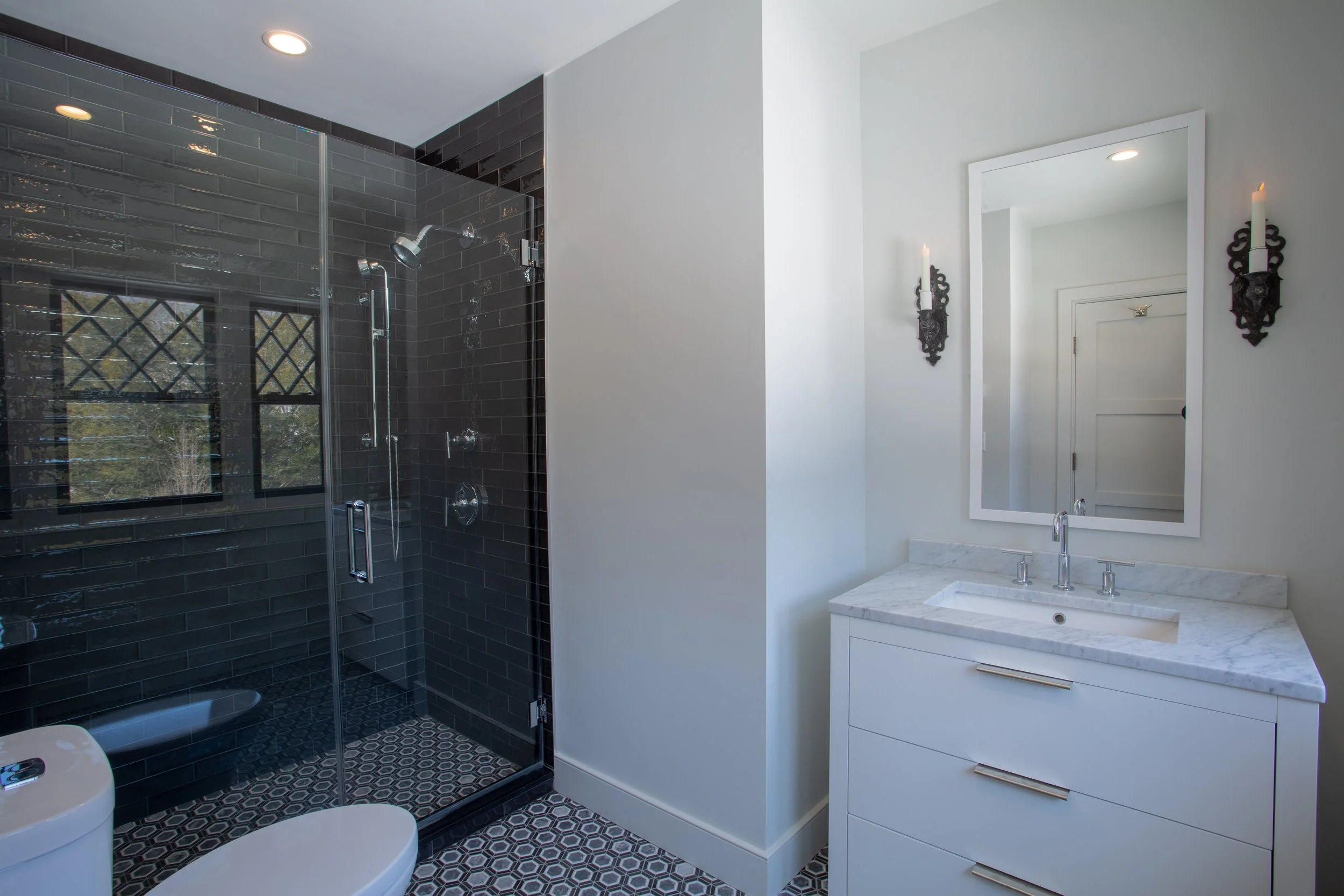 Modern bathroom with black-tiled walk-in shower, white vanity with marble countertop, large mirror, and decorative wall sconces.