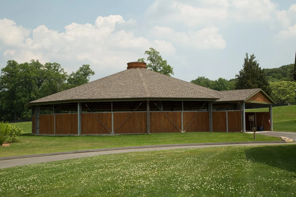 A large round wooden pavilion with a shingled conical roof, situated in a grassy park area with trees and a cloudy sky in the background.