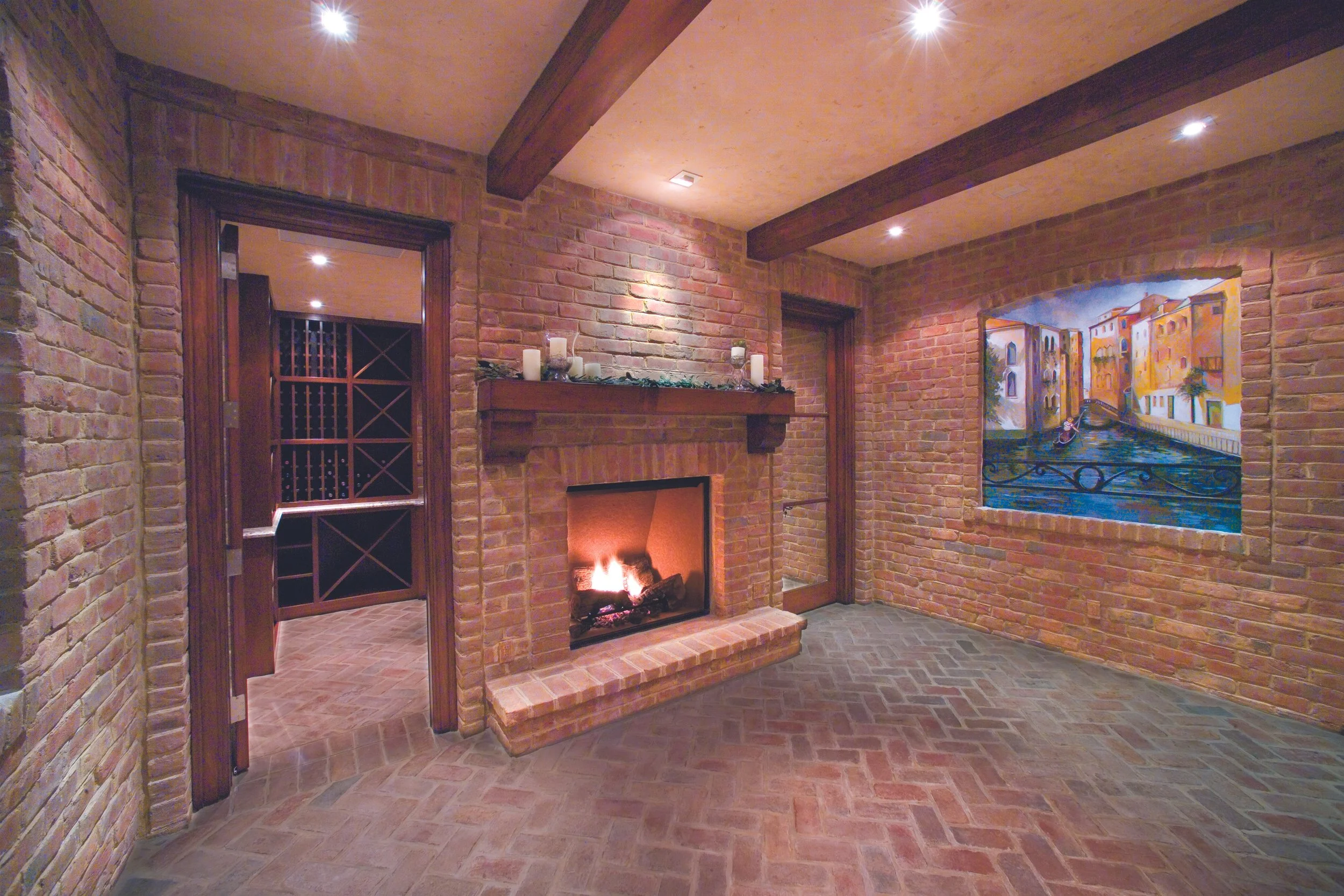 A cozy room with exposed brick walls, a brick fireplace with candles on the mantel, artwork of a canal scene on the wall, multiple ceiling lights, and a wood beam ceiling.