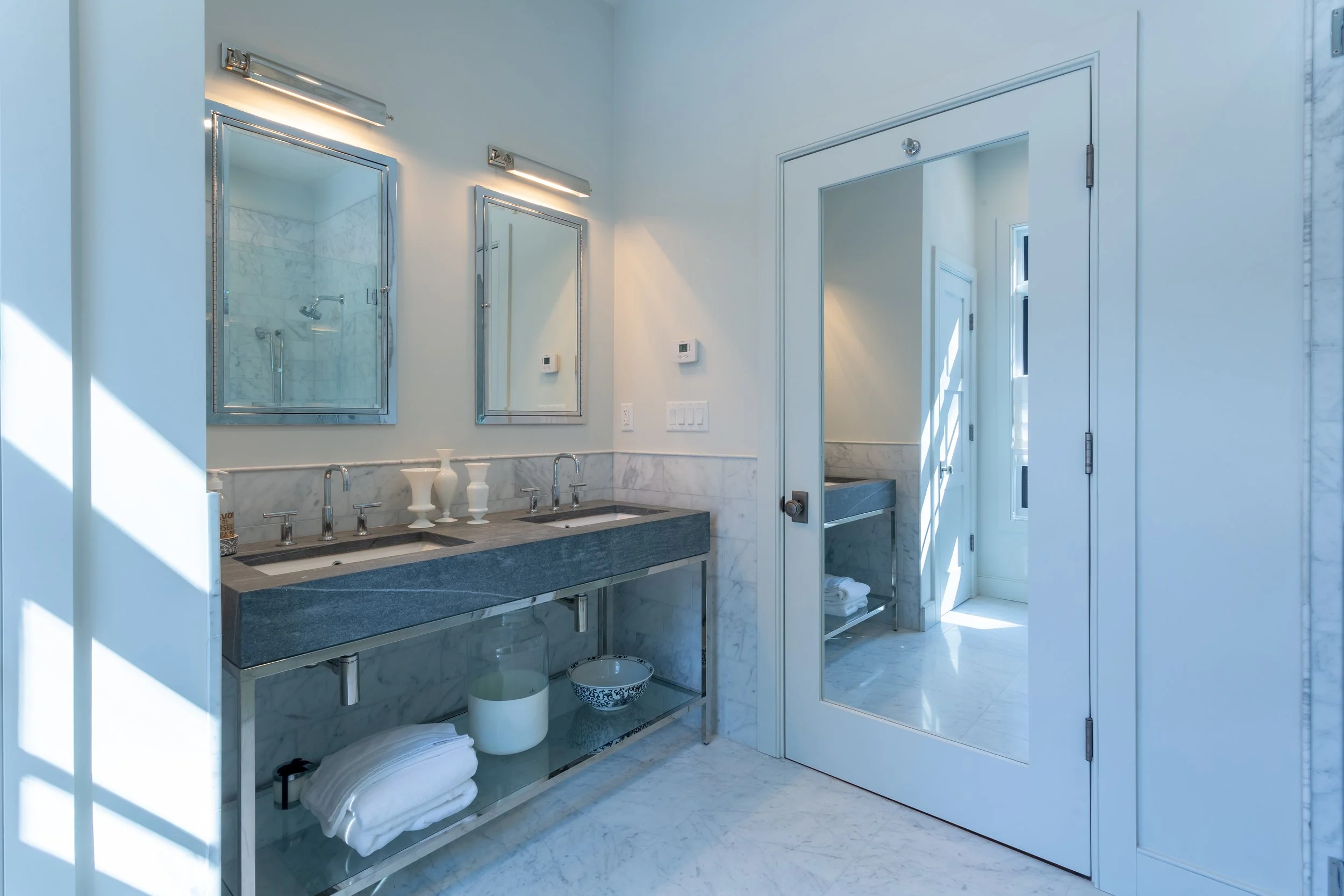 Modern bathroom with double sinks, mirrors, and a glass shower door, illuminated by wall-mounted lights.