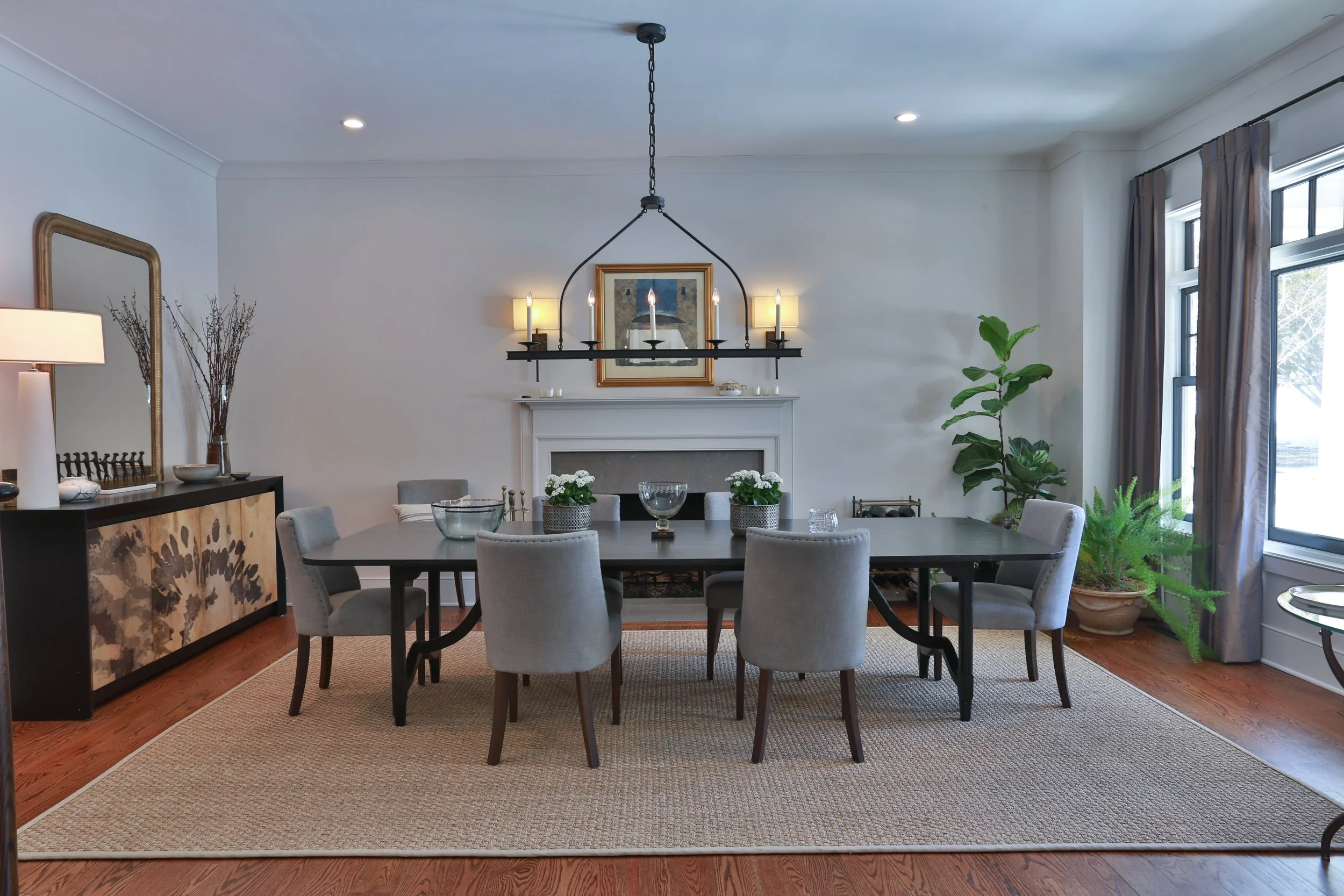 A dining room with a rectangular table and eight gray chairs, a fireplace with a mantle, a chandelier with candles, large windows with curtains, and indoor plants.